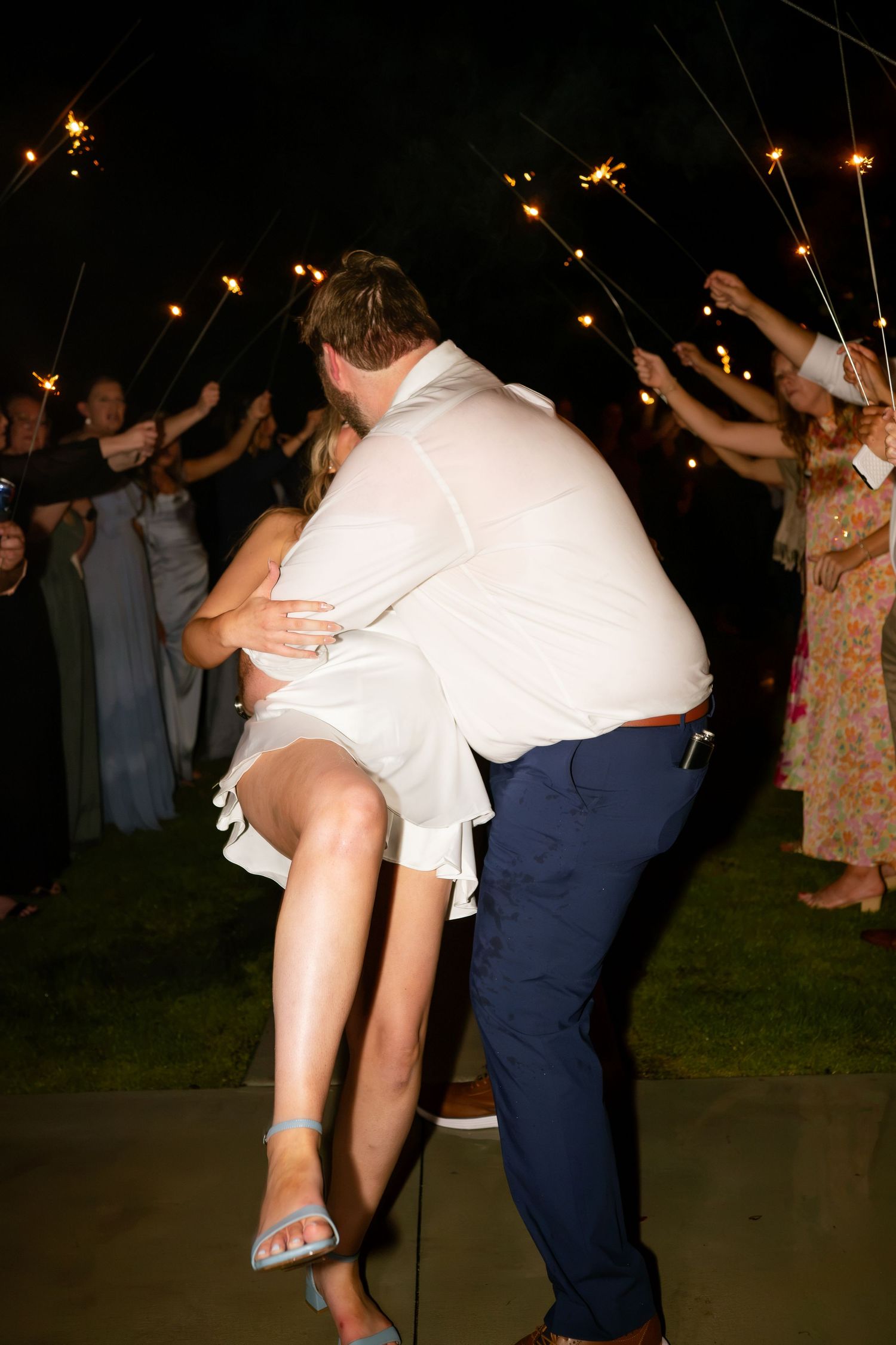 A groom lifts his bride in a romantic dip during a sparkler send-off exit at their outdoor evening wedding reception.
