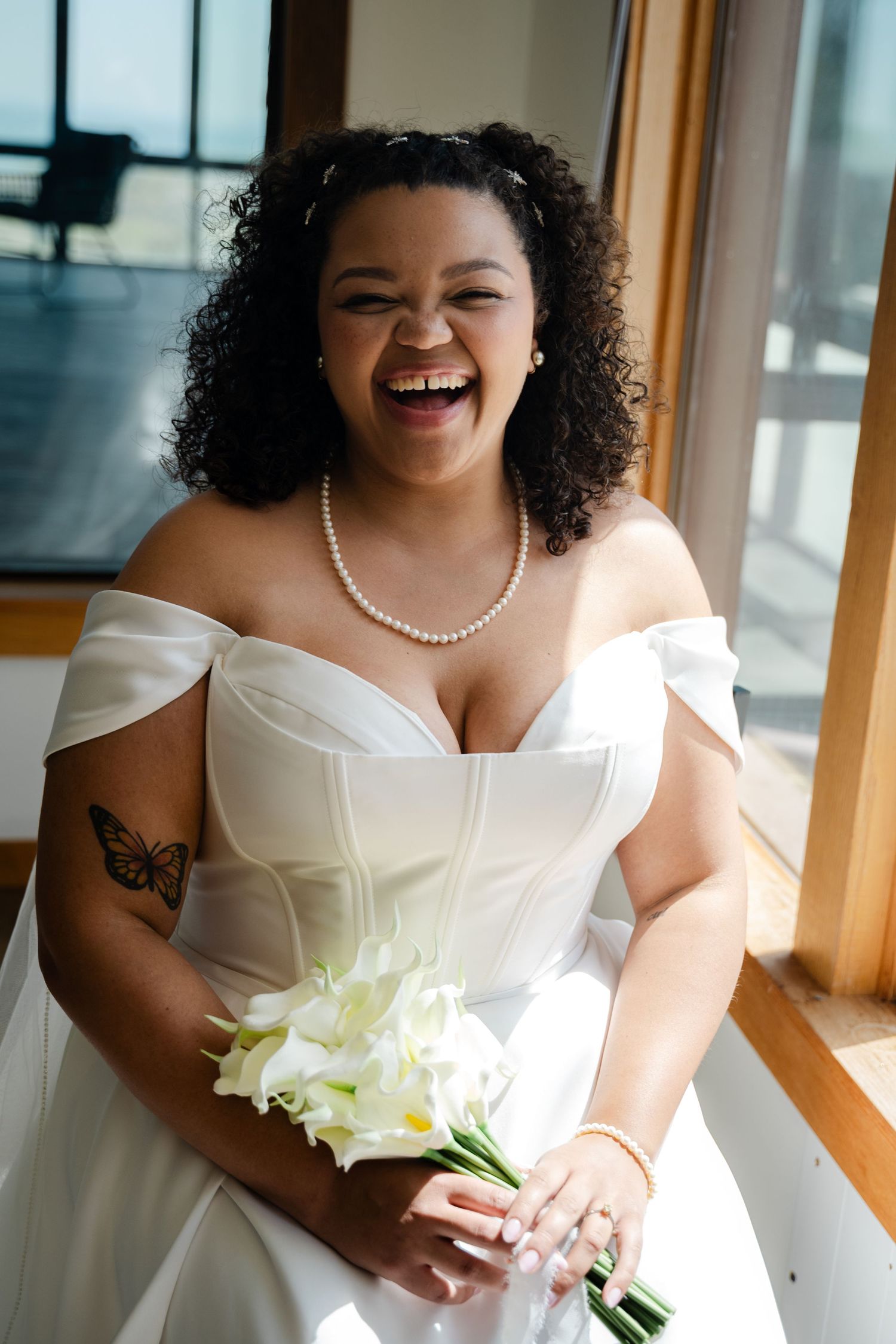 Joyful bride in off-shoulder white gown laughing while holding white calla lily bouquet.