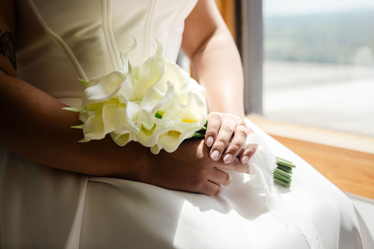Bride in white gown holding elegant white calla lily bouquet near sunlit window.