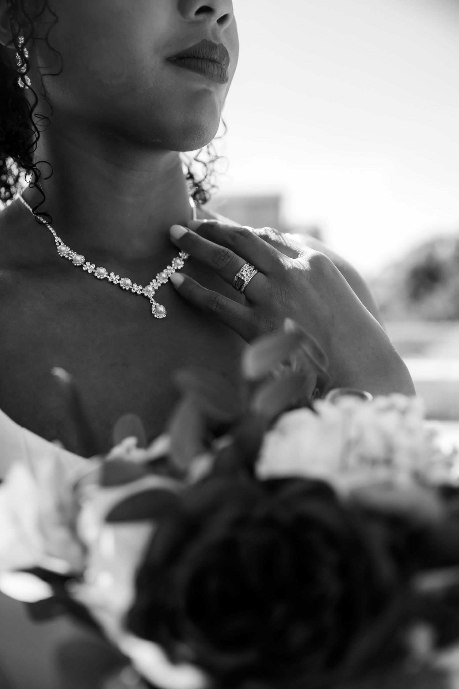 Black and white close-up of bride touching diamond necklace while holding bouquet with roses.