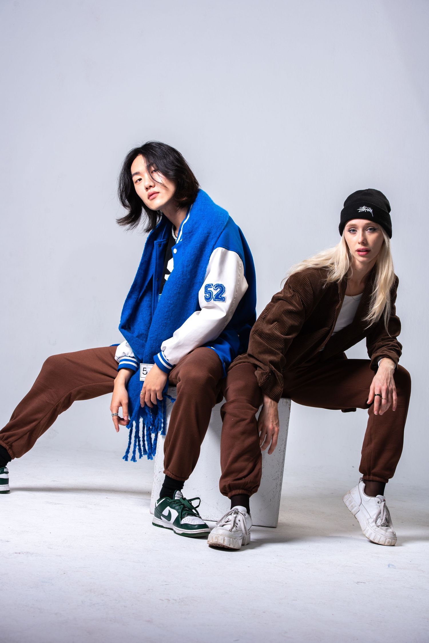 Two stylish women pose in casual streetwear with varsity jacket and brown outfits in a studio.