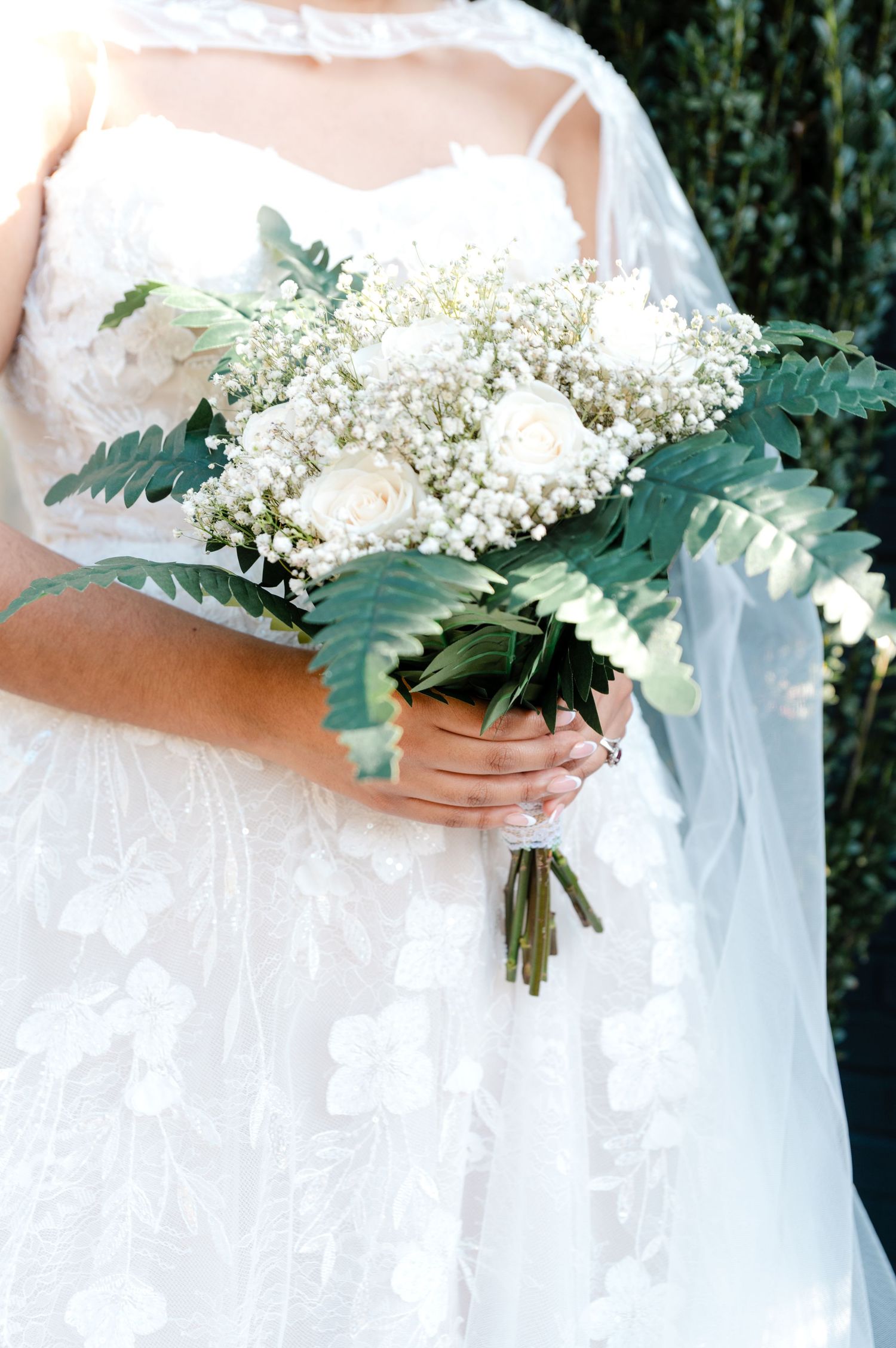 Bride in lace wedding gown holding white bouquet with roses, baby's breath, and green ferns.