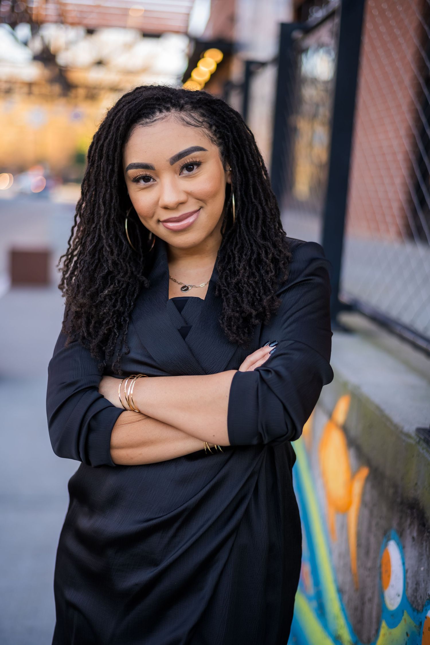 A confident young Black woman with locs poses arms crossed in a black blazer against an urban graffiti wall.