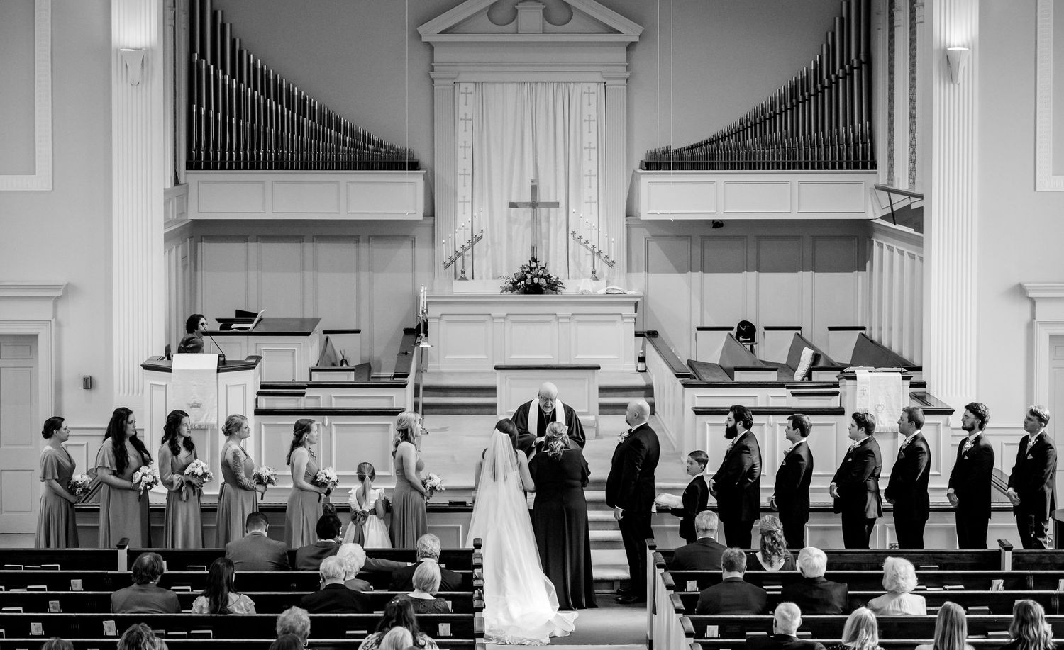 Black and white wide shot of wedding ceremony in grand church with full bridal party at altar.