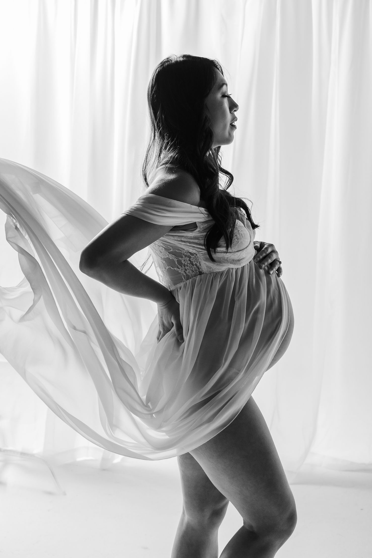 Black and white silhouette of pregnant woman in flowing sheer fabric near bright curtained window.