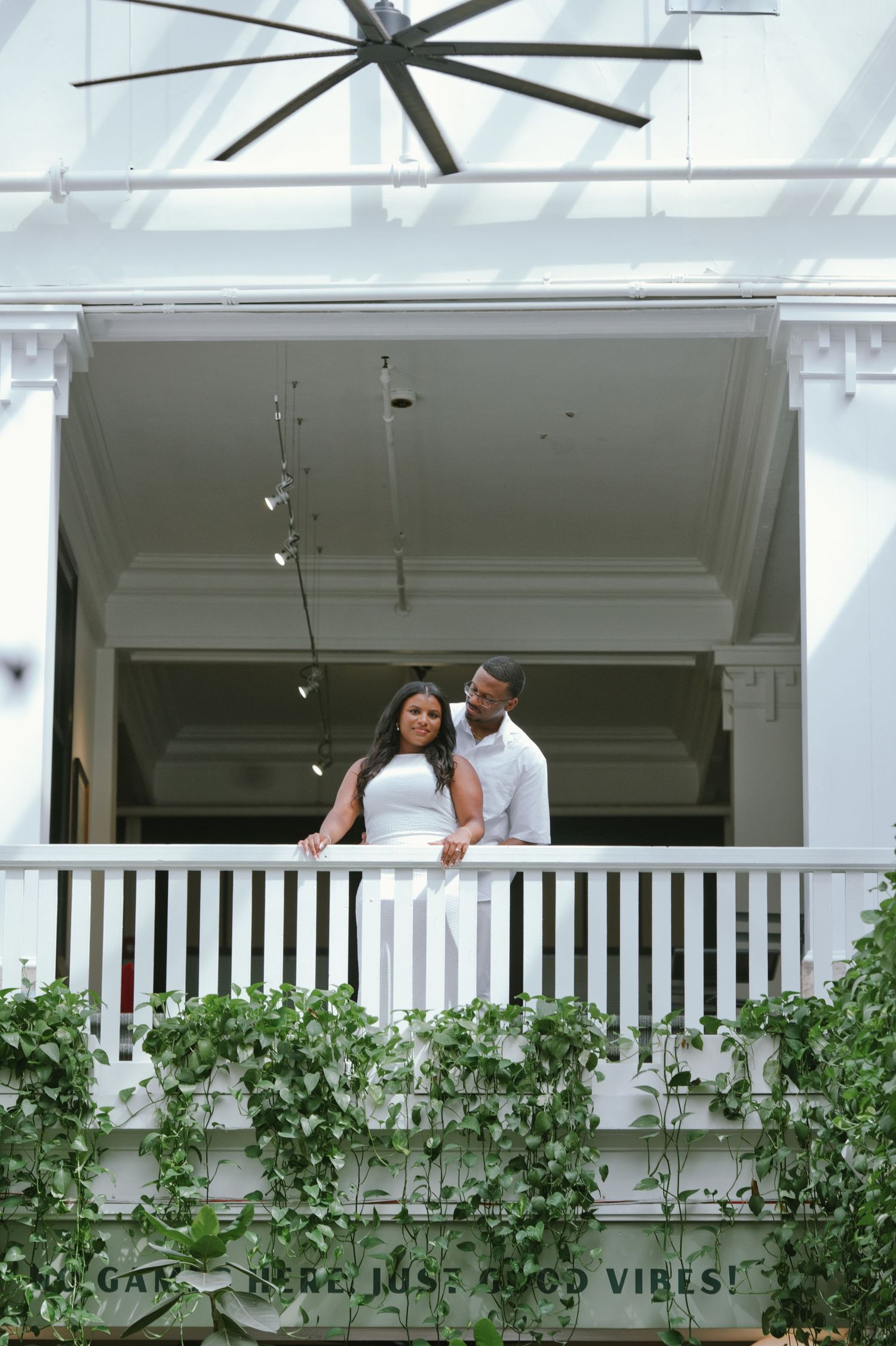 Elegant Black couple in white attire standing together on a vine-covered balcony at wedding venue.