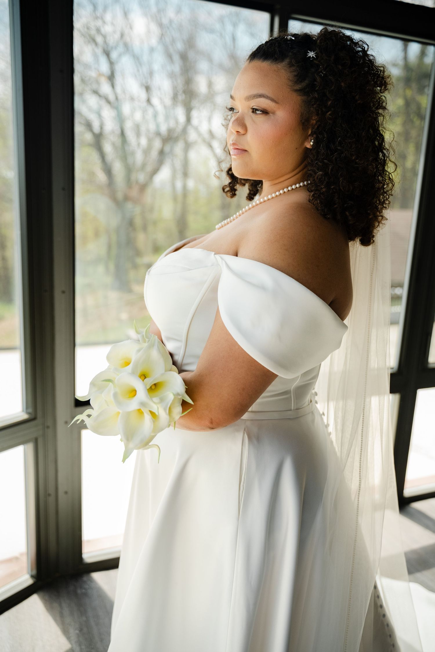 Bride in off-shoulder white gown holding calla lily bouquet gazing out large windows.