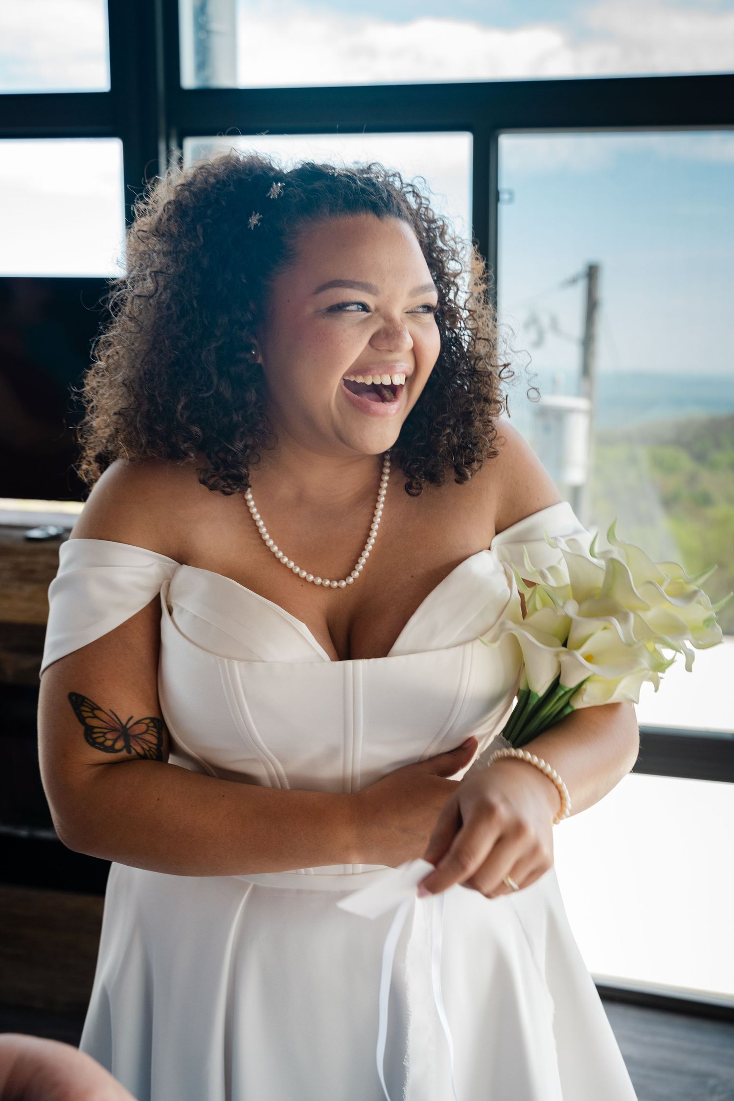 Laughing bride in off-shoulder white dress holding calla lilies with butterfly arm tattoo.