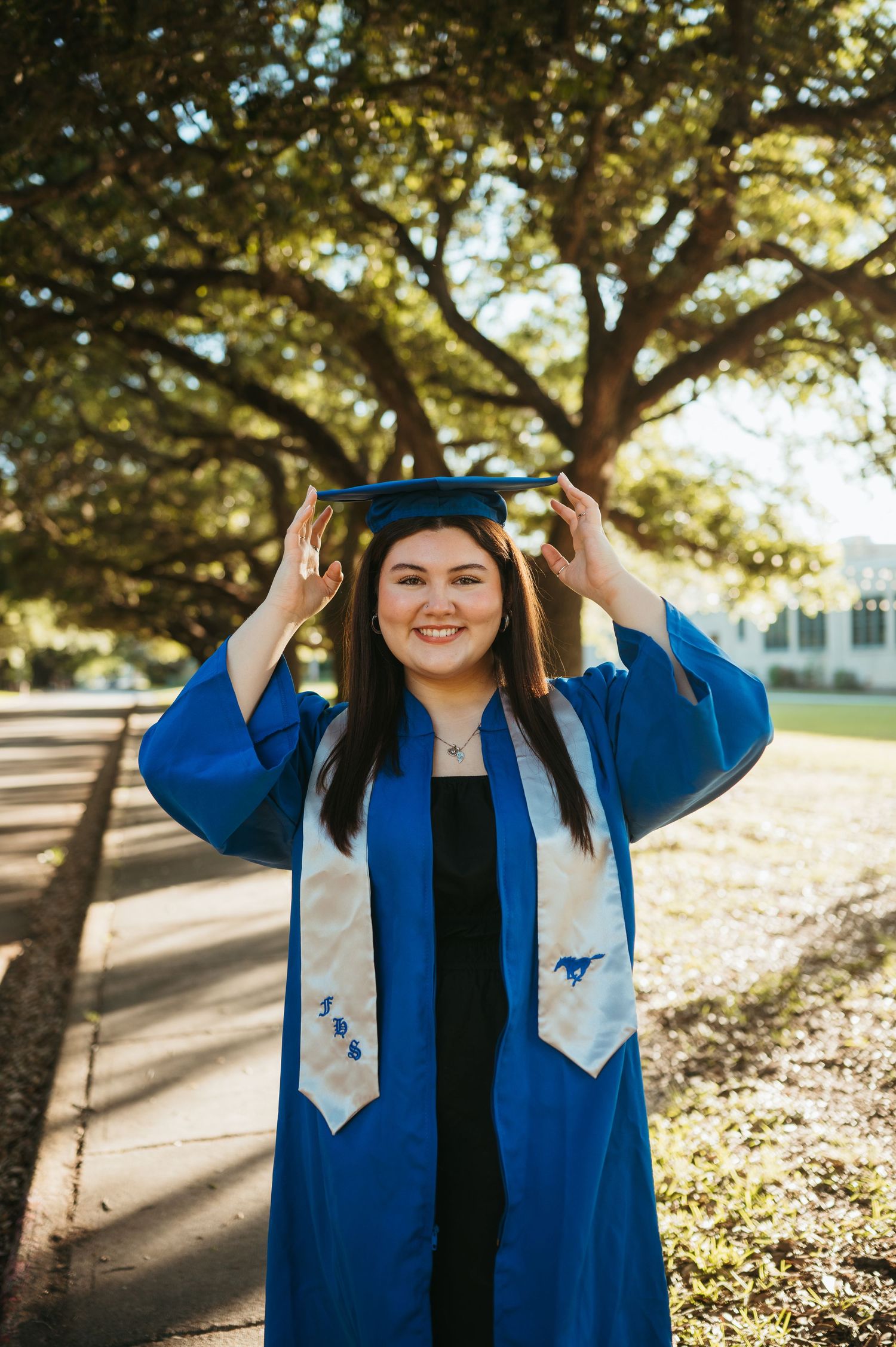 A senior dons her cap and gown during her senior session among the oak trees