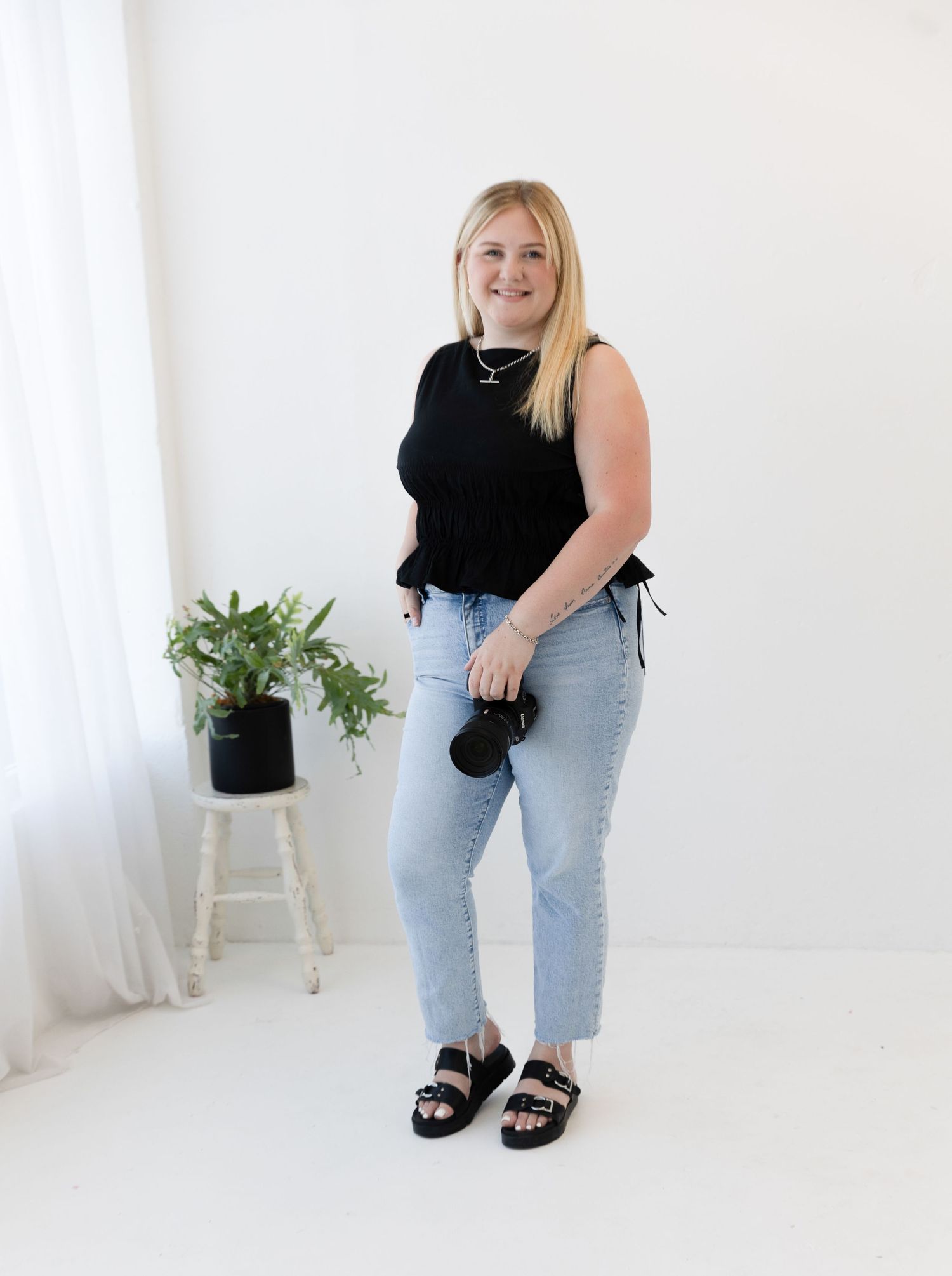 Casual fashion look with black top, light jeans and sandals against white background with potted plant.