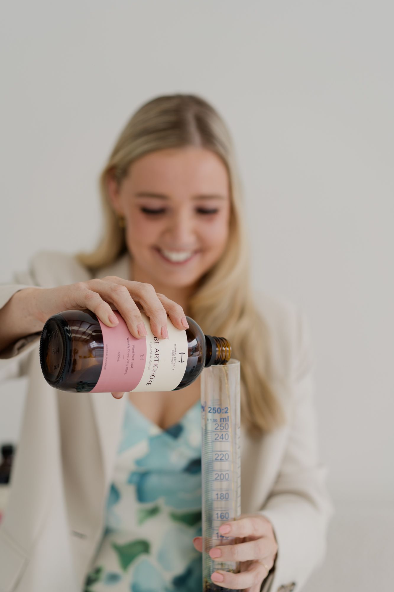 Blonde woman pouring liquid supplement into measuring cylinder from pink-labeled brown bottle.
