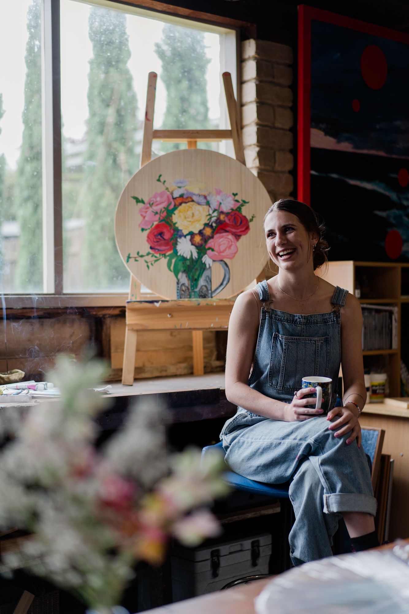 Smiling female artist in denim overalls sitting in her art studio holding a mug near floral painting.