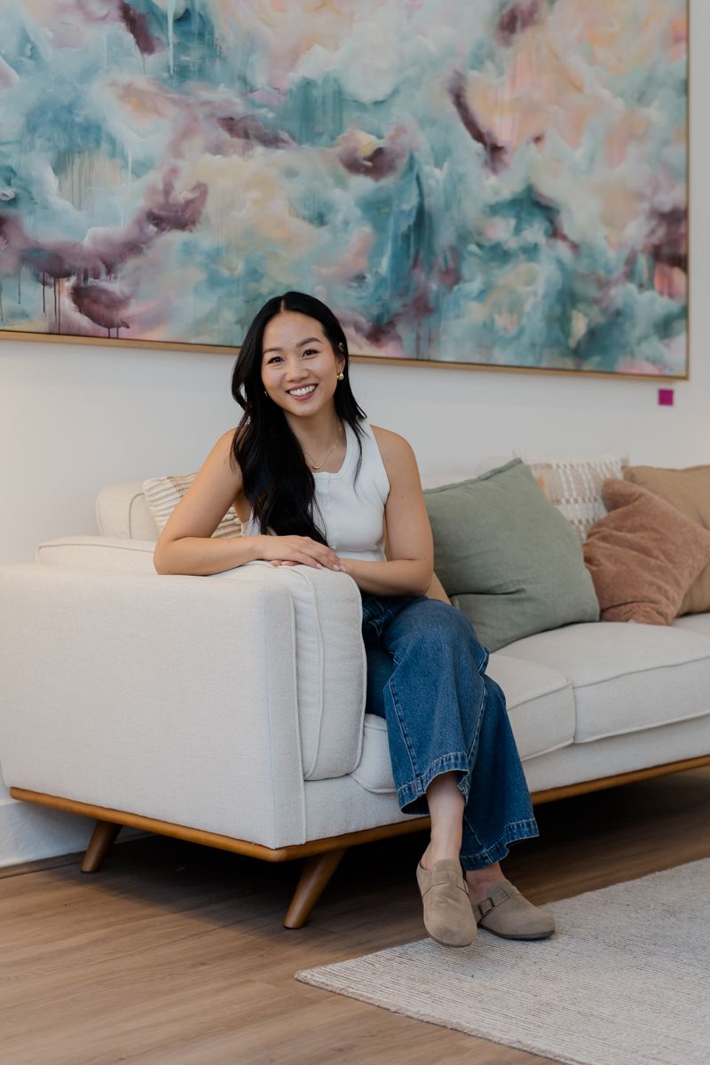 Smiling woman sitting on white sofa with colorful abstract painting on wall behind her.