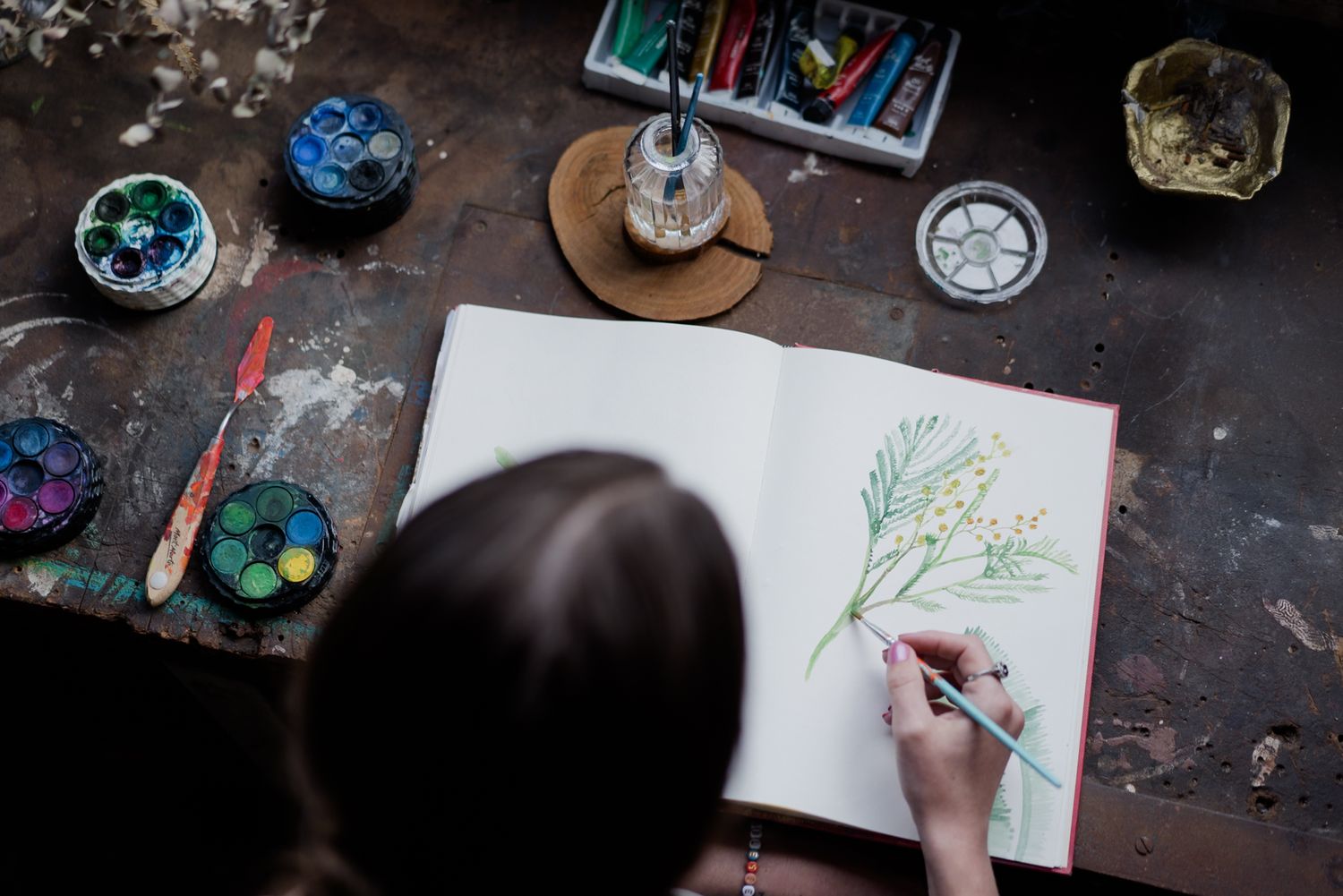 Overhead view of artist sketching botanical illustration in sketchbook surrounded by watercolor paints.