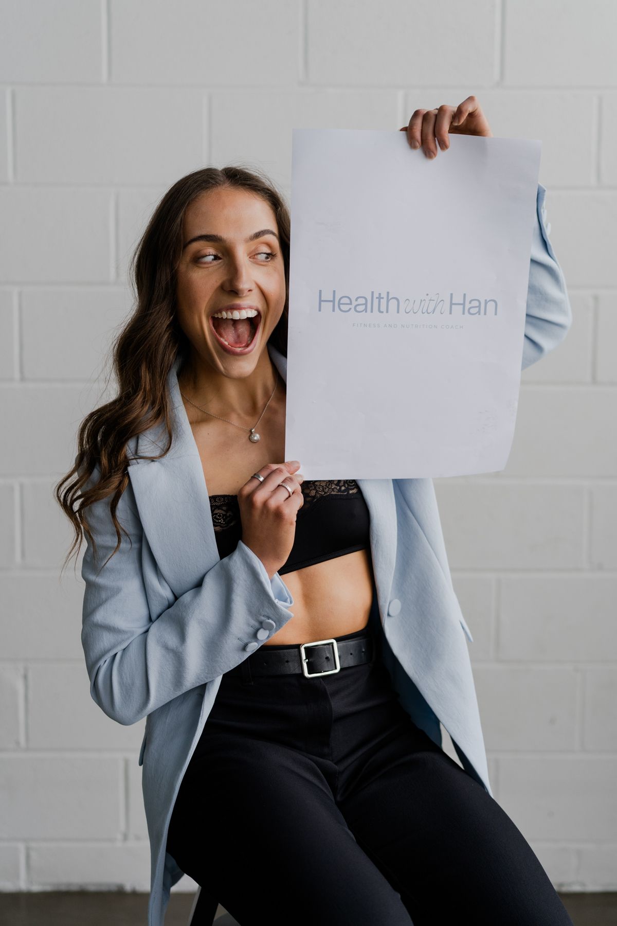Excited woman in light blue blazer holding Health with Han logo paper with open mouth expression against white wall.