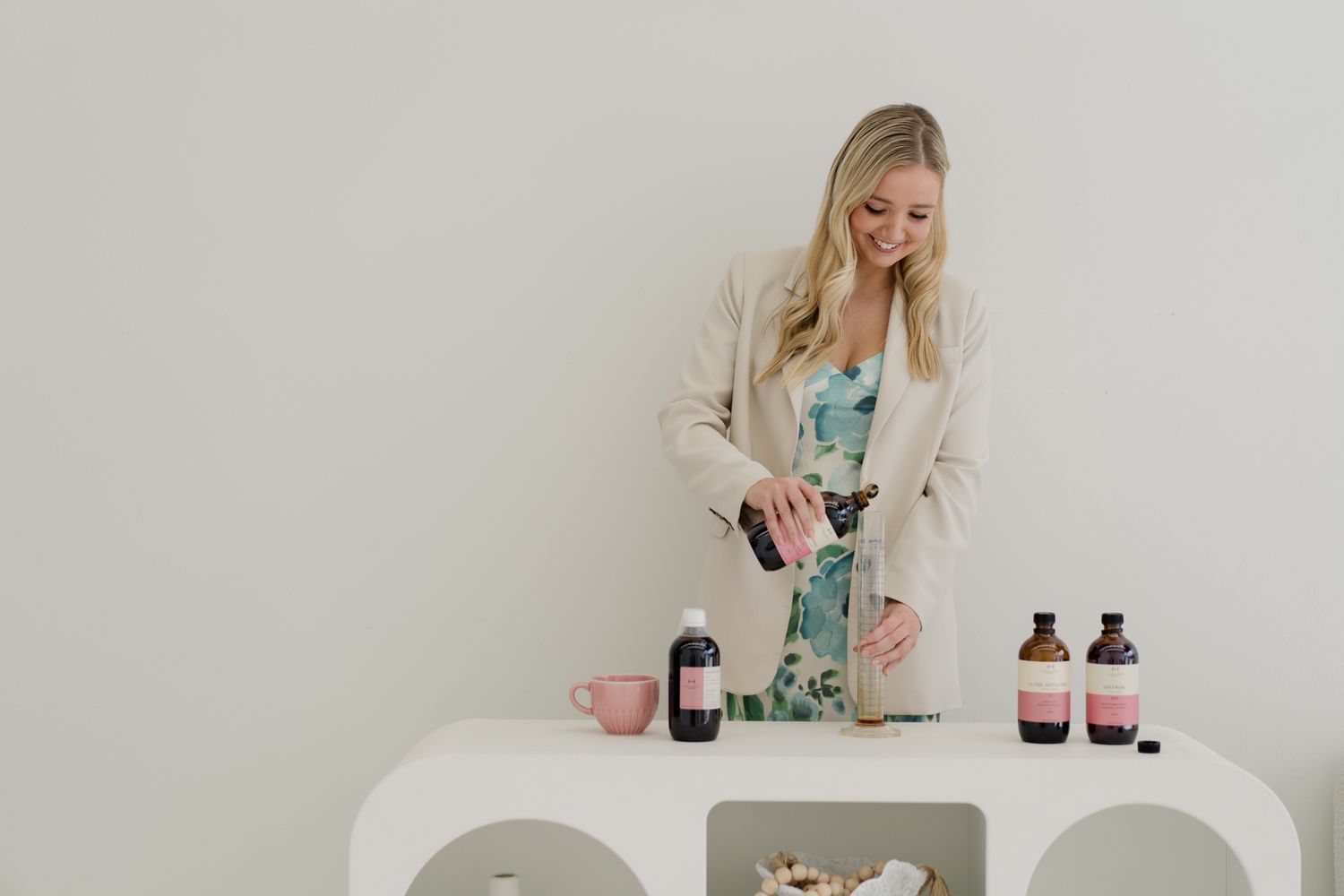 Blonde woman in floral dress and blazer arranging health supplement bottles on white table.
