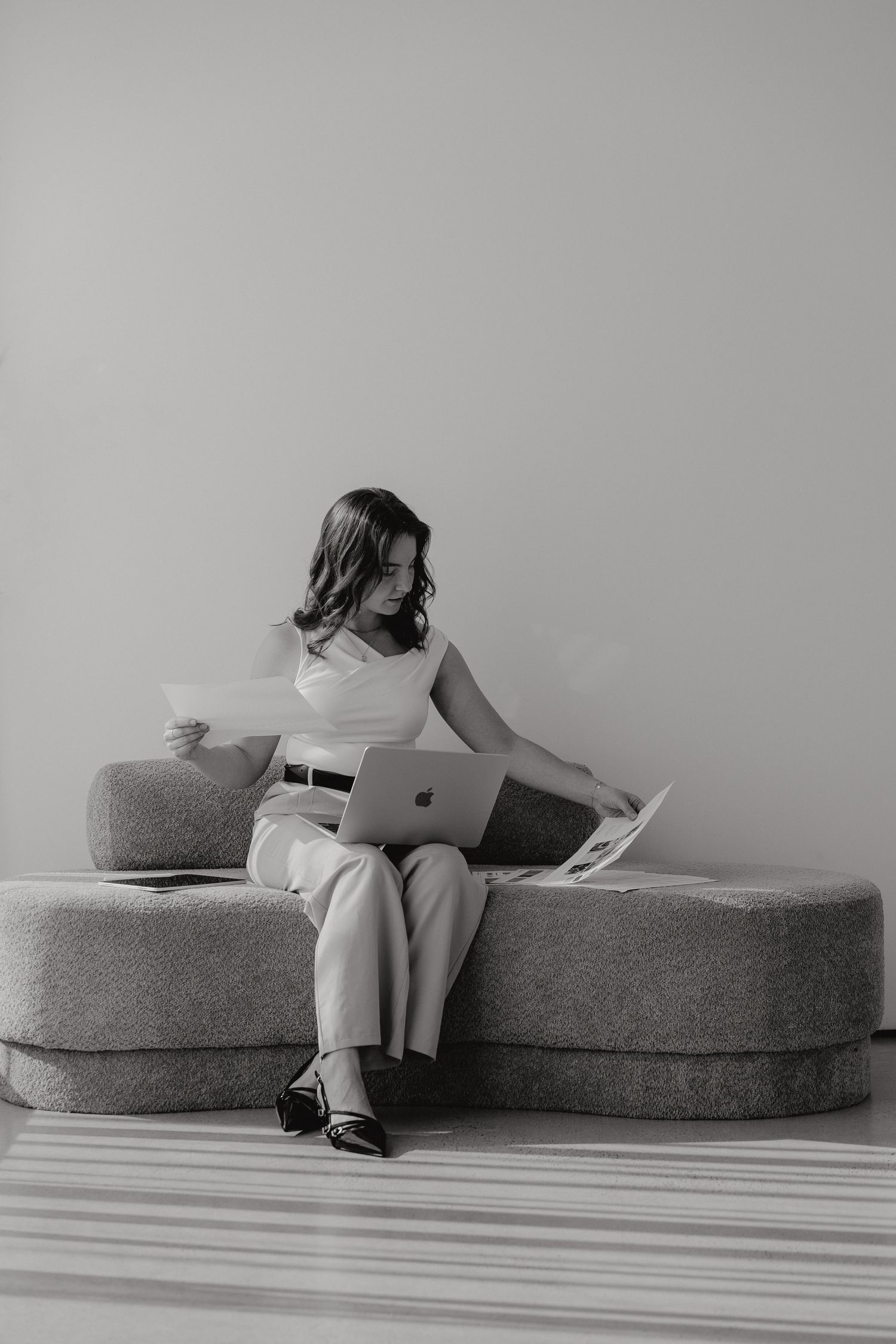 Woman in white outfit reviewing documents while working on laptop on modern sofa.
