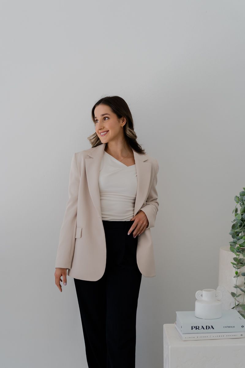 Smiling woman in beige blazer and black trousers posing against white wall near Prada book.