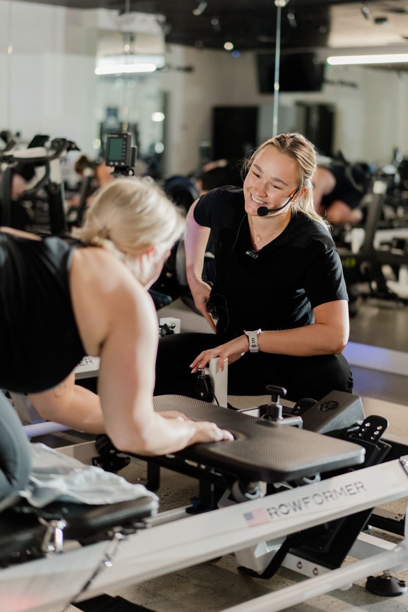 Fitness instructor with headset assists a client on a Rowformer machine during a gym class.