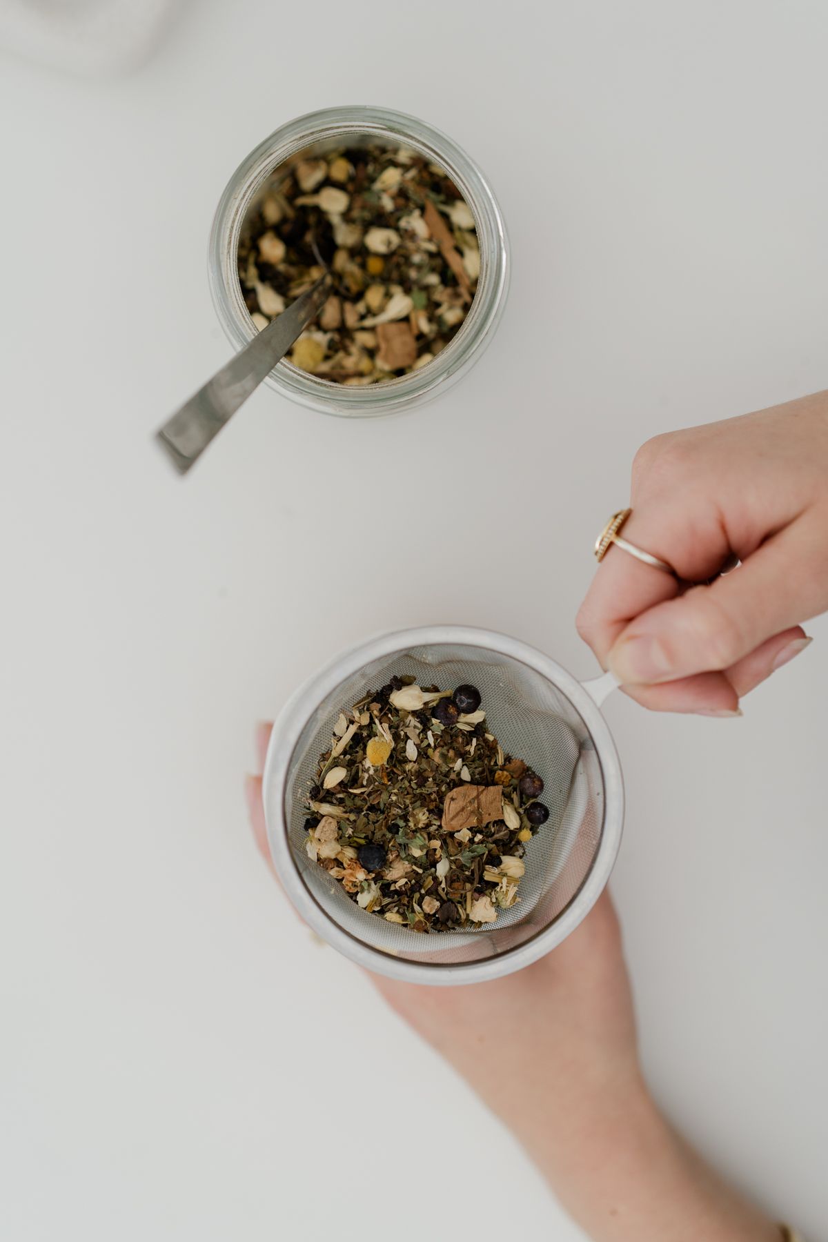 Overhead view of hands holding a mesh tea strainer filled with loose leaf herbal tea beside a glass jar.