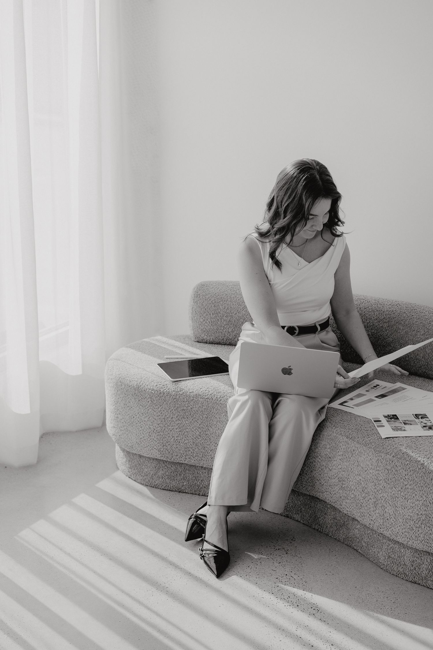 Woman in white sleeveless top working on MacBook laptop while reviewing documents on sofa.