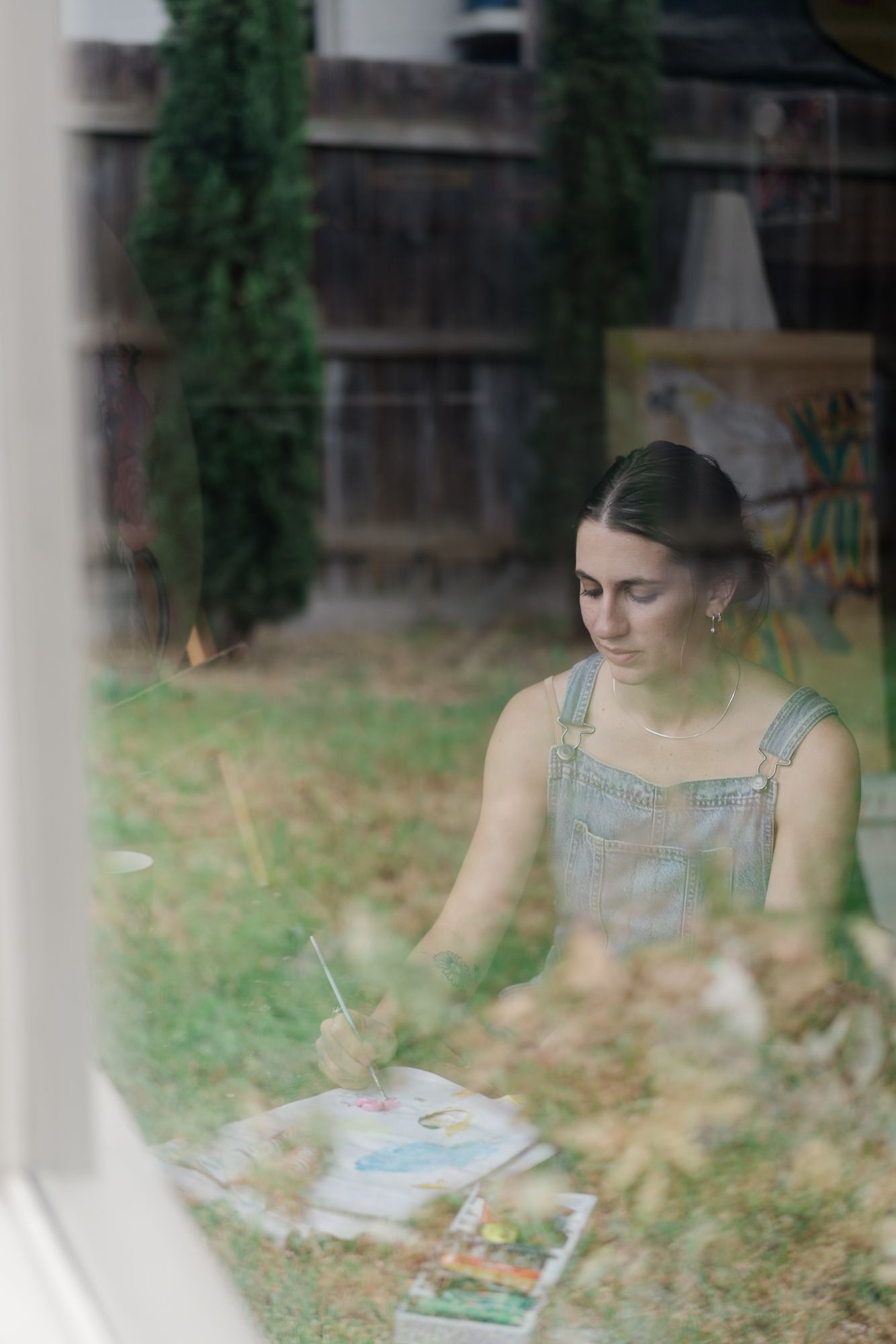 Female artist in denim overalls painting watercolors outdoors in a garden, viewed through a window.