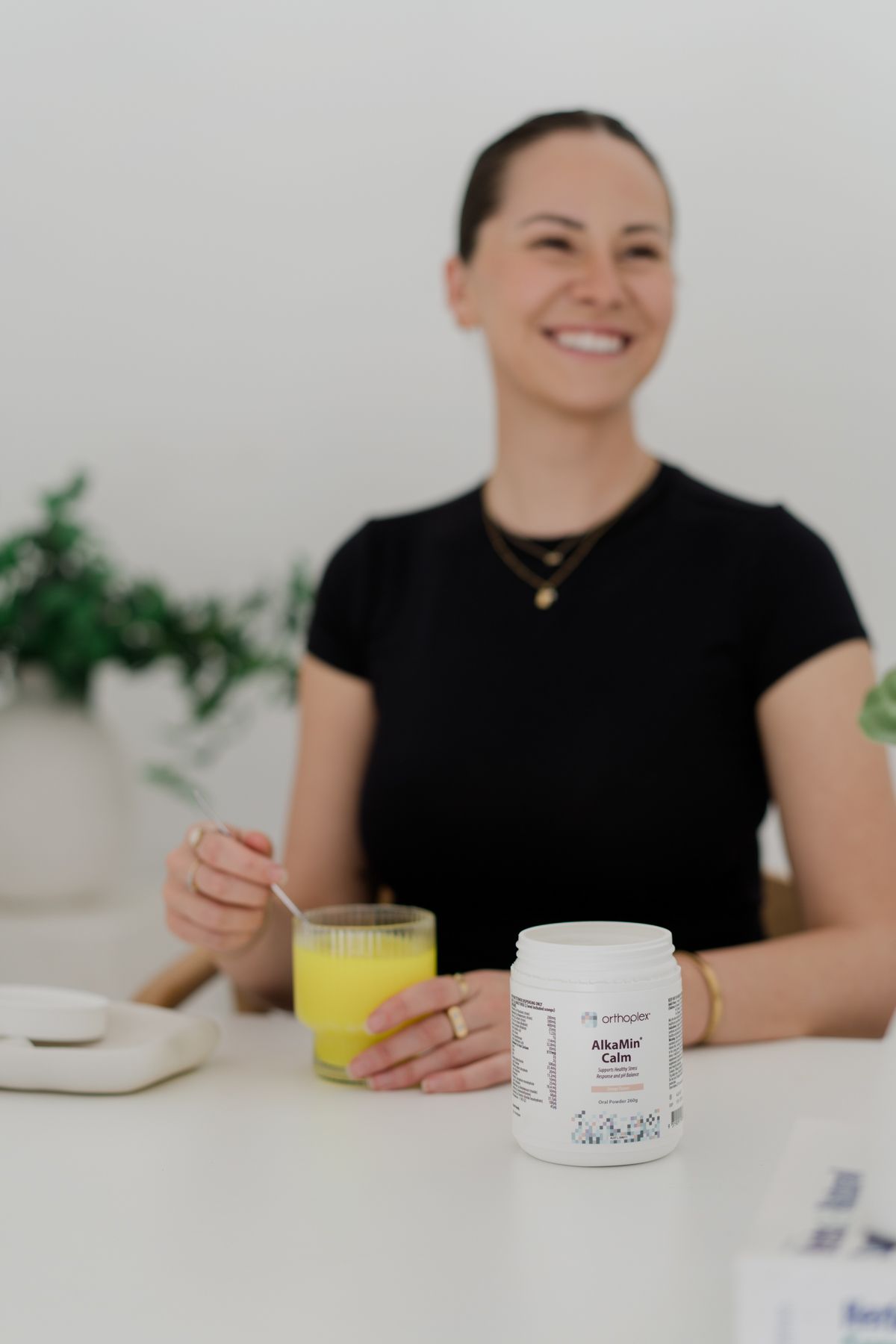 Smiling woman in black top with AlkaMin Calm supplement jar and yellow drink at table.