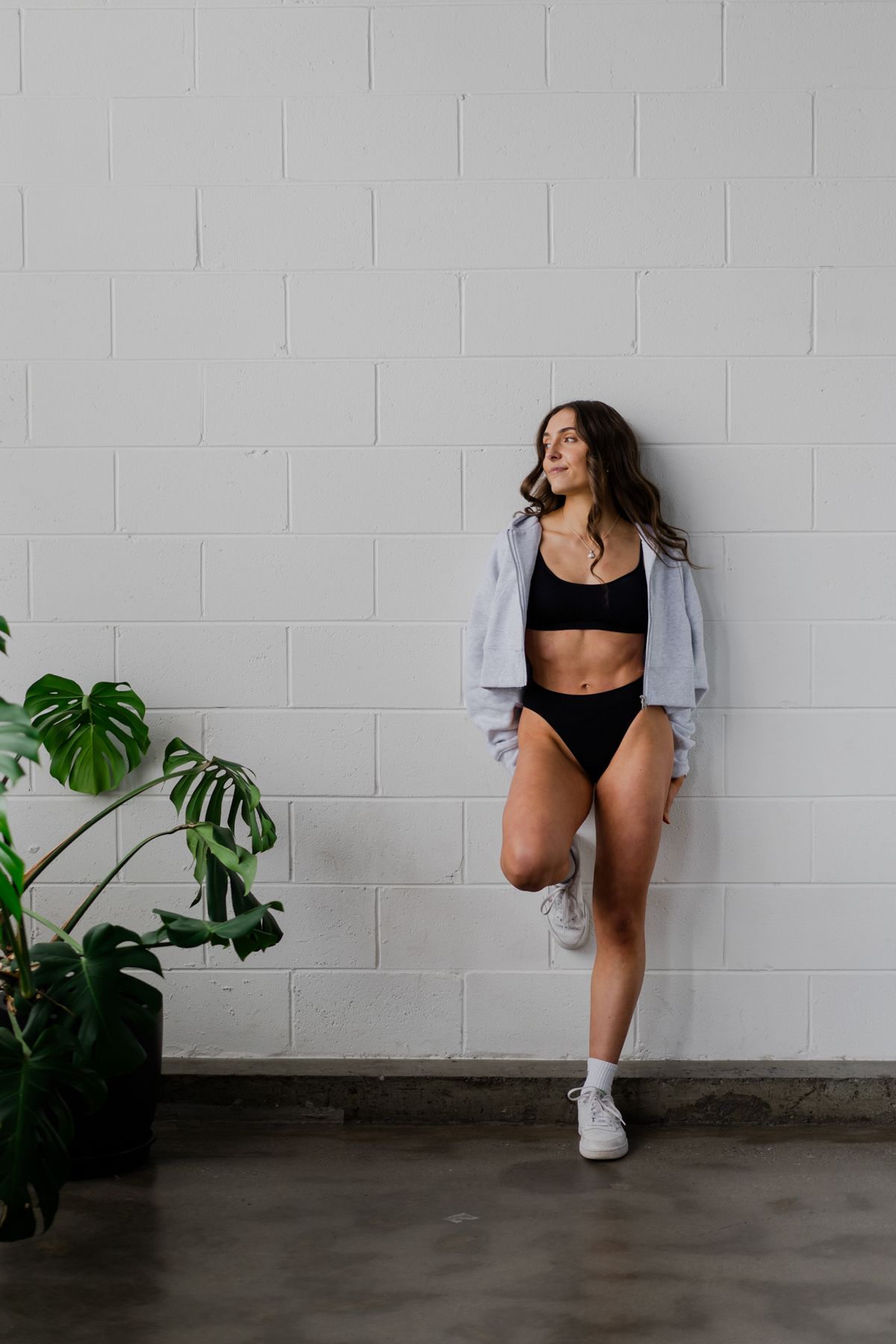 Athletic woman in black sports bra and shorts leaning against a white brick wall beside a monstera plant.