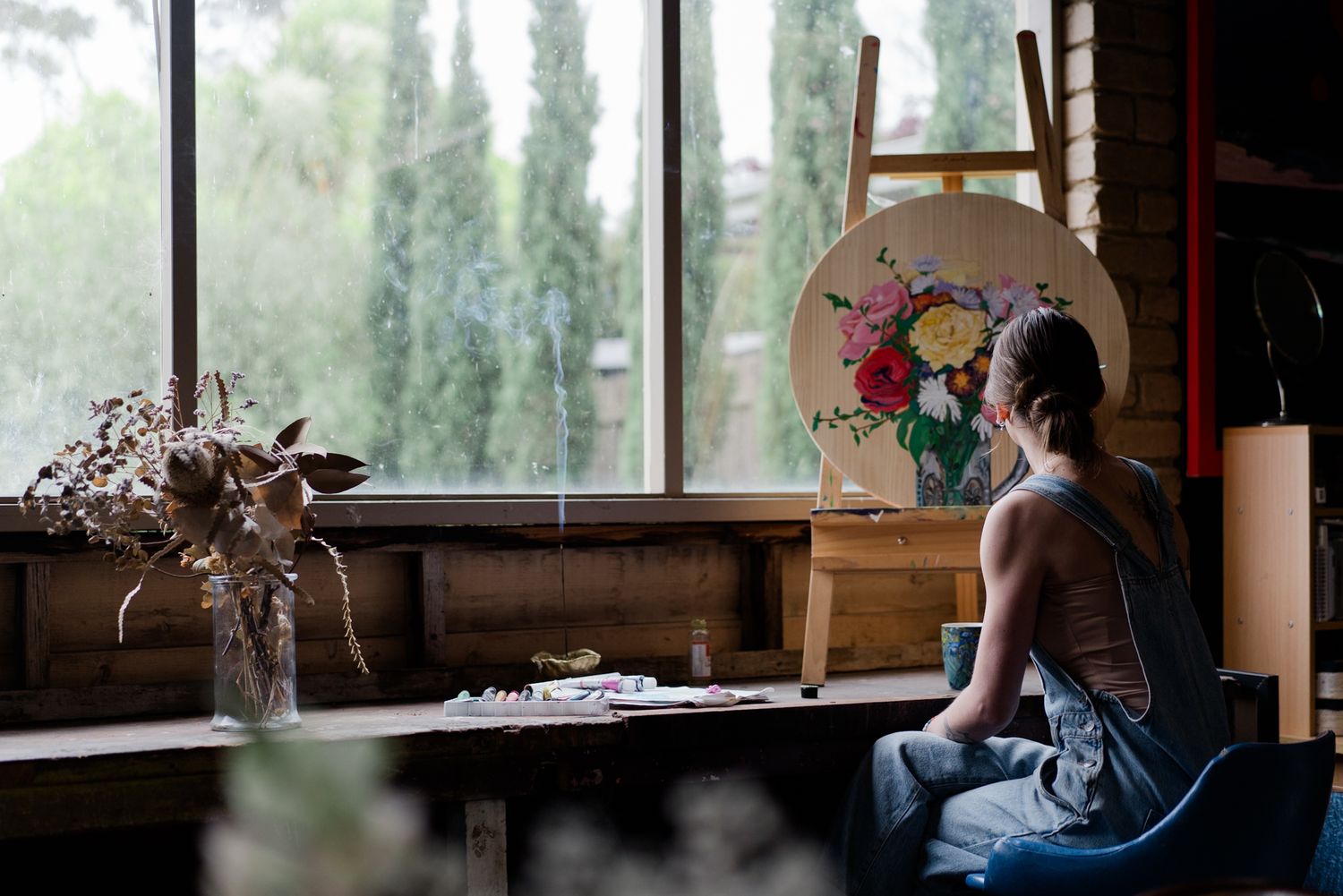 Female artist in denim overalls sitting before a round floral painting on easel in a rustic studio.