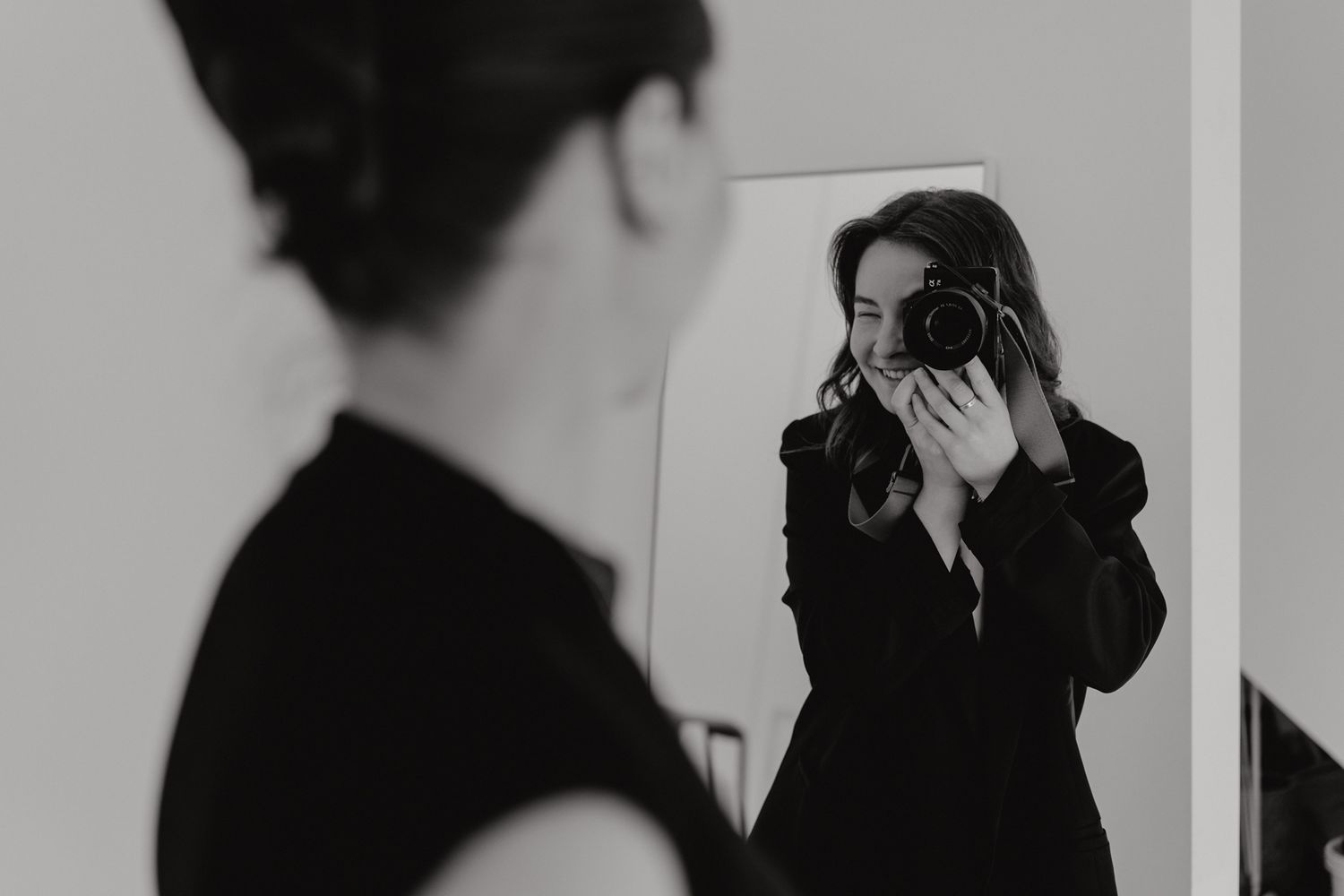 Female photographer smiling while taking mirror selfie with DSLR camera in black outfit.