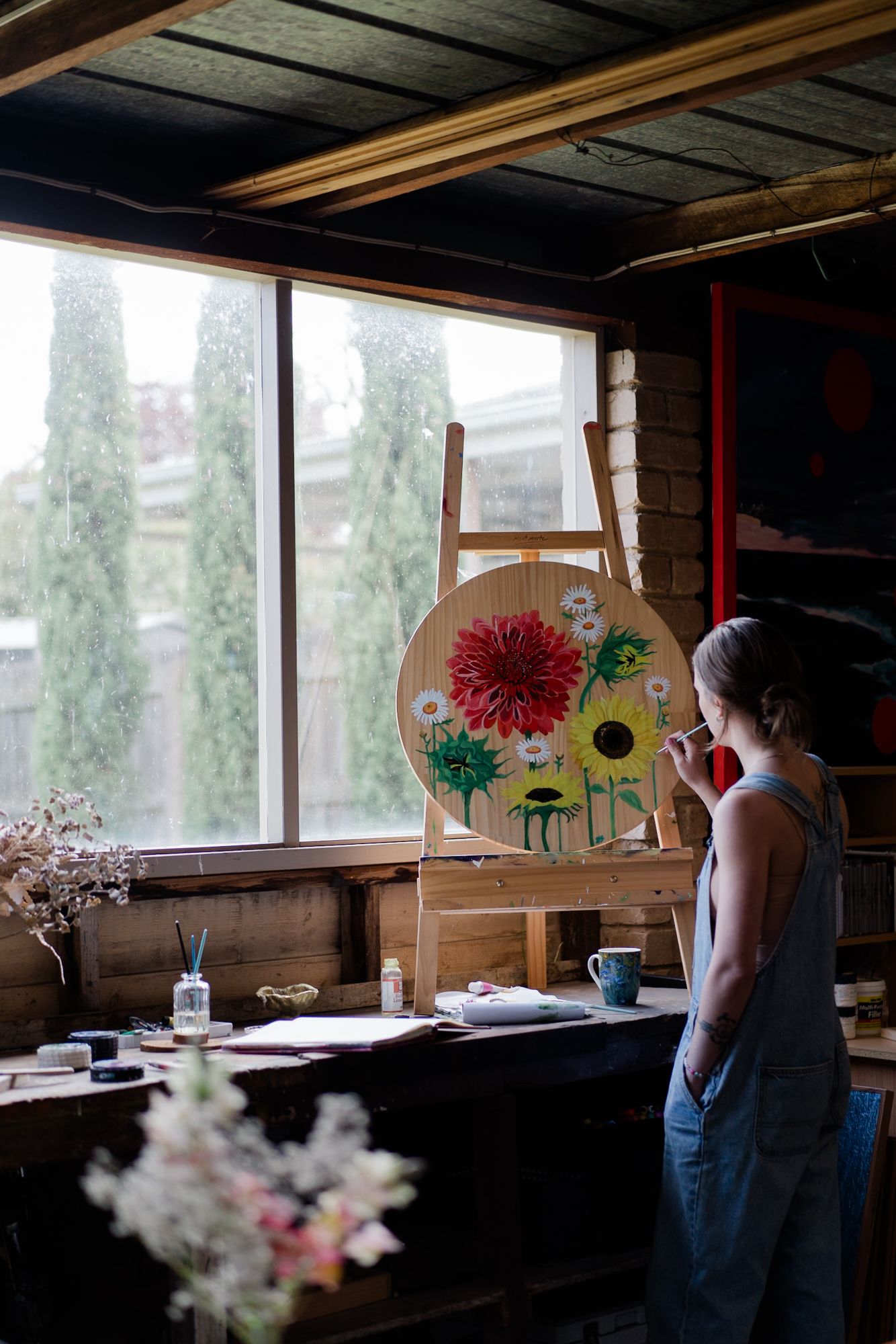 Female artist in denim overalls painting colorful flowers on a round canvas in a rustic studio.