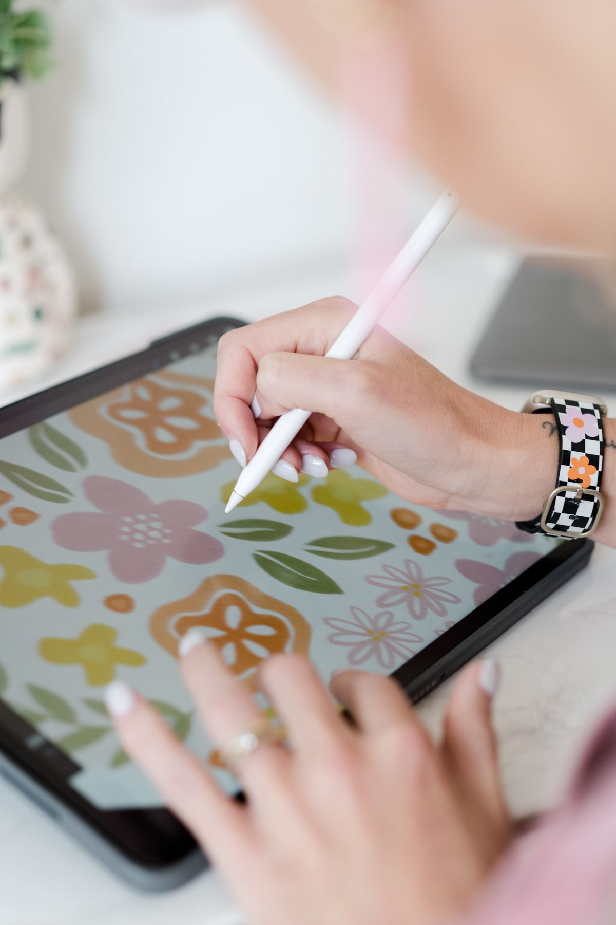 Close-up of hands using Apple Pencil to draw colorful floral pattern on tablet with checkered band.