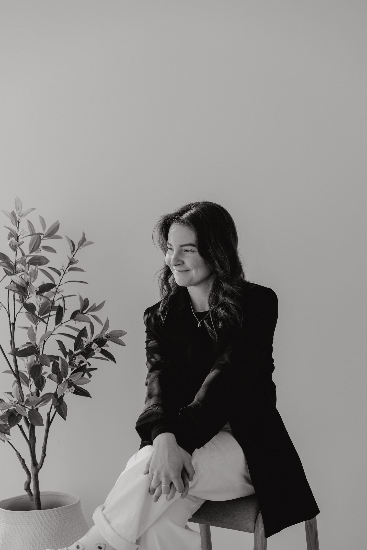 Young woman in black blazer sitting on stool next to potted plant in black and white photo.
