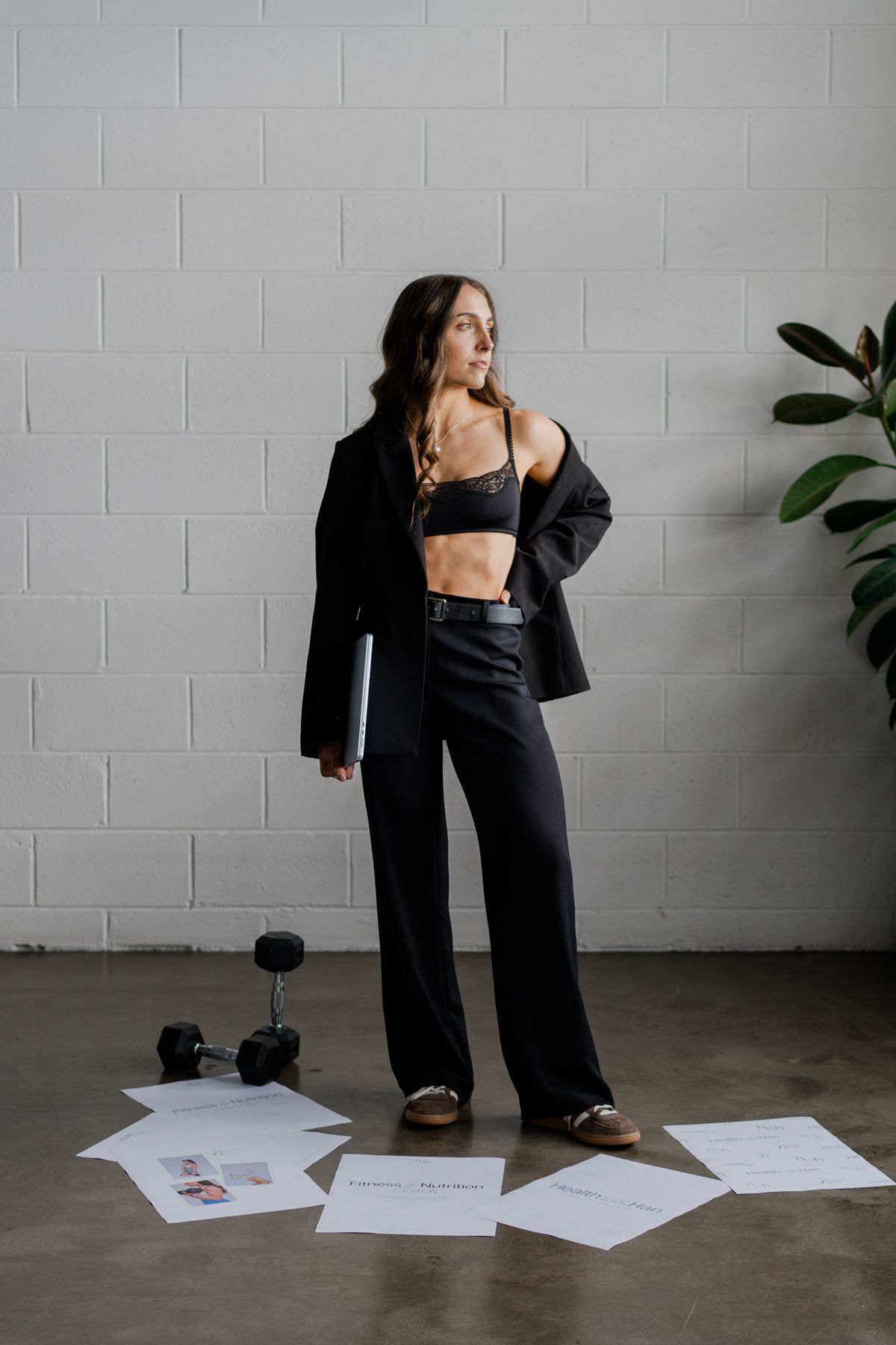 Fitness coach in black outfit poses with laptop among scattered nutrition program papers and dumbbells on concrete floor.