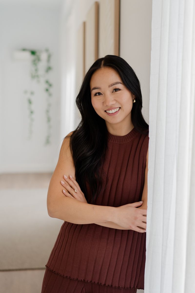 Smiling Asian woman in burgundy pleated top leans against white column with arms crossed indoors.