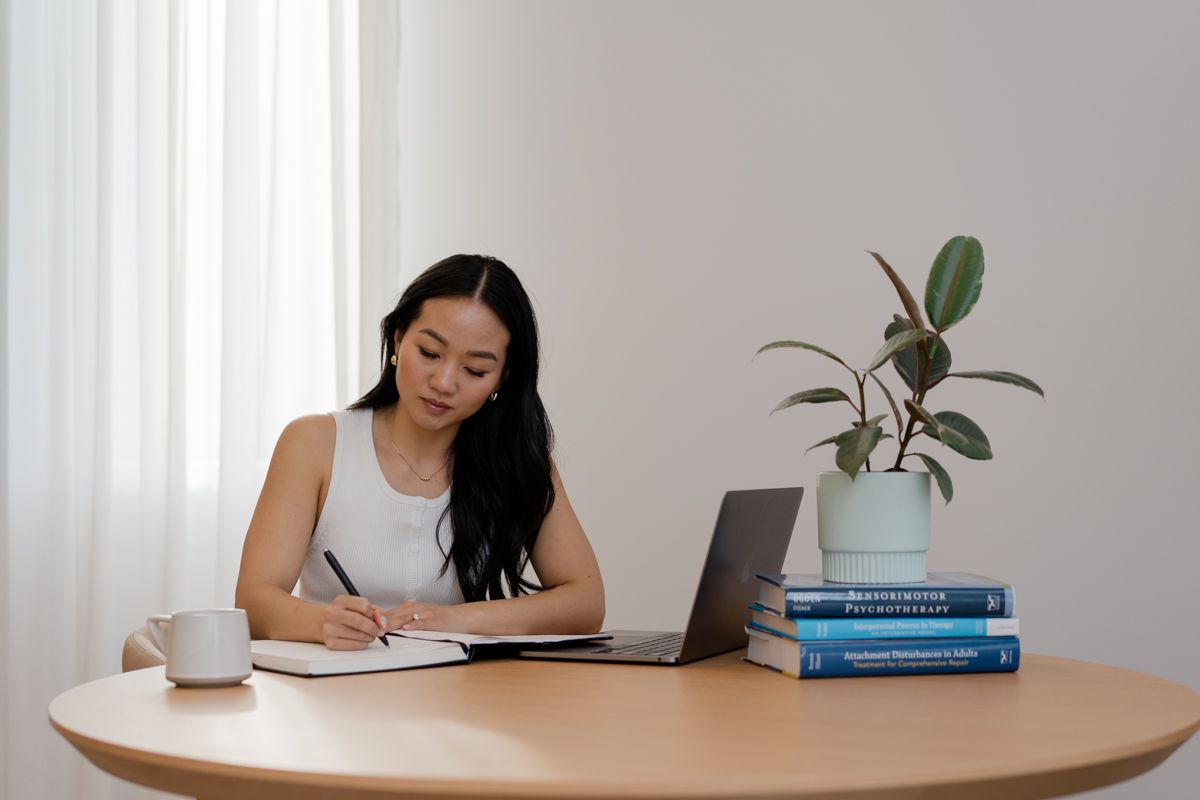Woman in white top writing in notebook at round desk with laptop and plant nearby.
