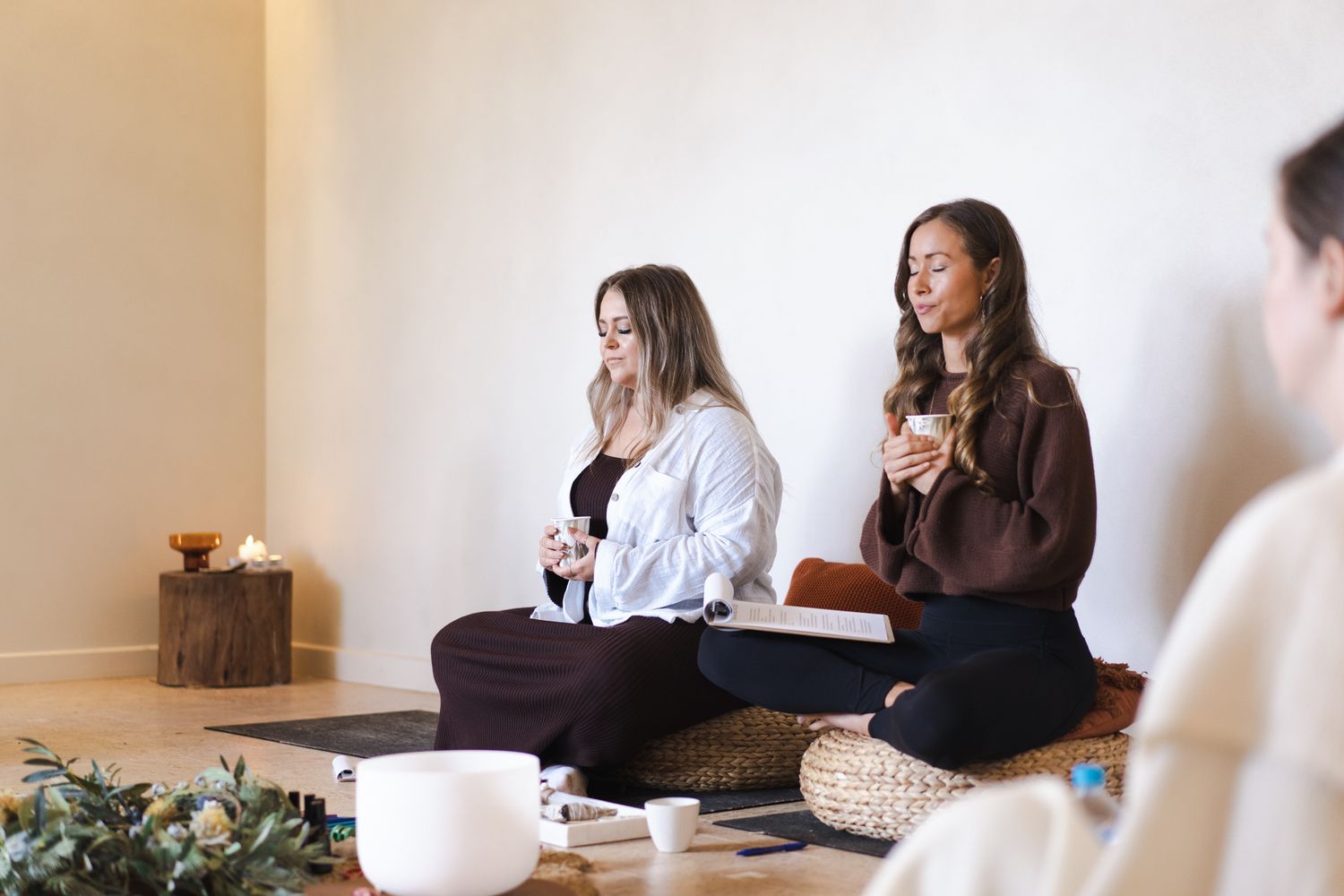 Two women sitting cross-legged on yoga mats in a bright studio, holding cups during a meditation session.