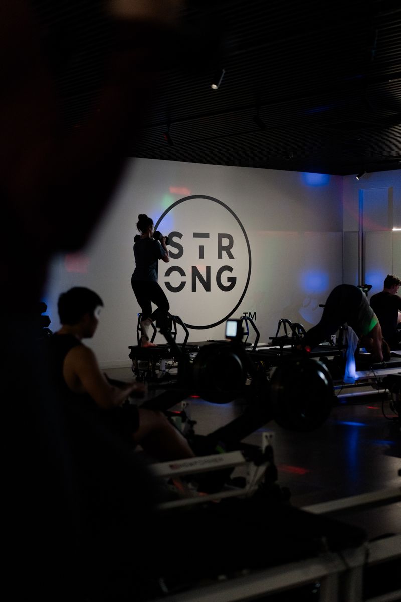 Silhouette of instructor on bike in front of illuminated STRONG logo during a dark fitness class.