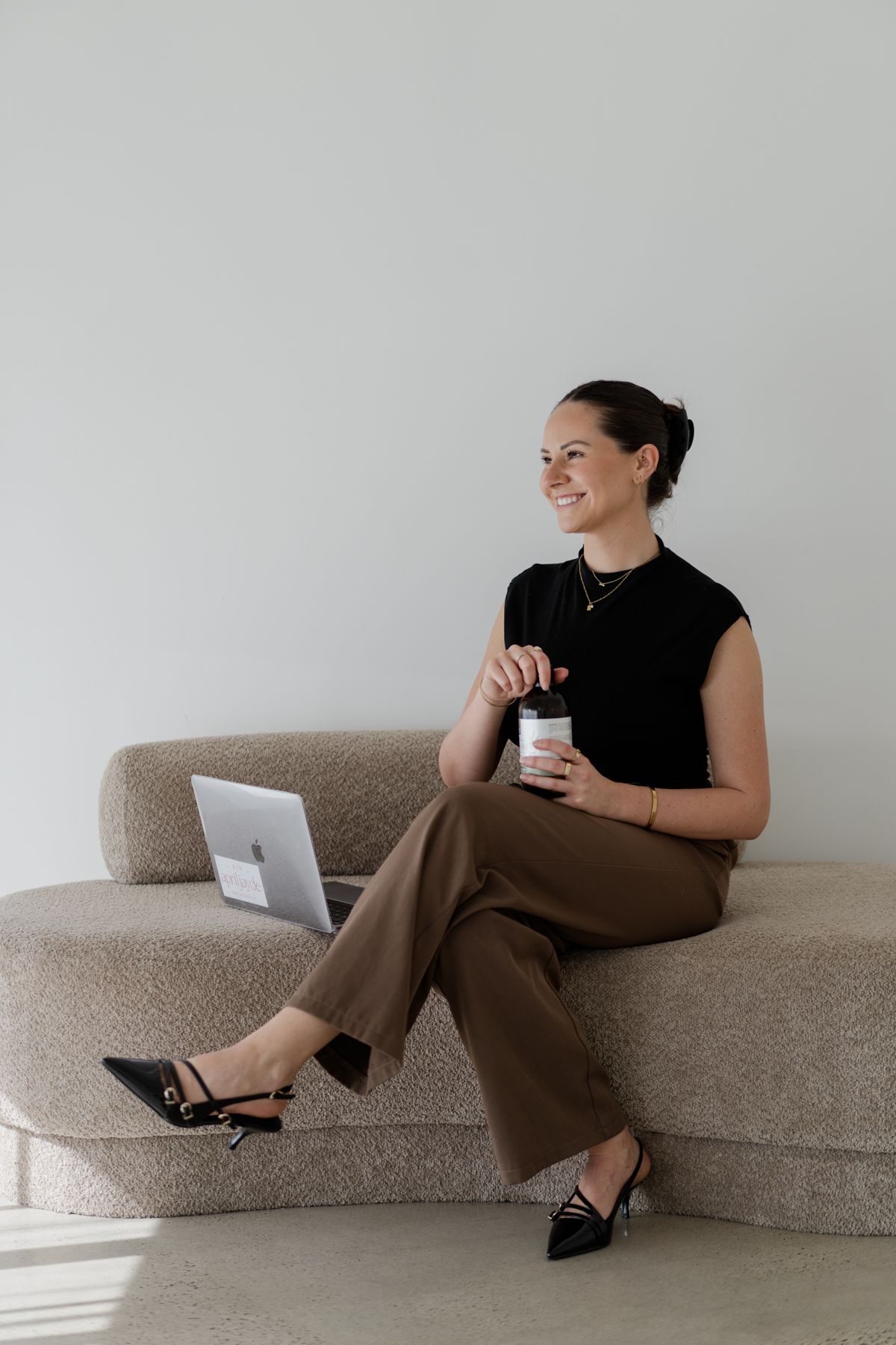 Smiling woman in black top and brown trousers sits on a couch holding a supplement jar beside a laptop.