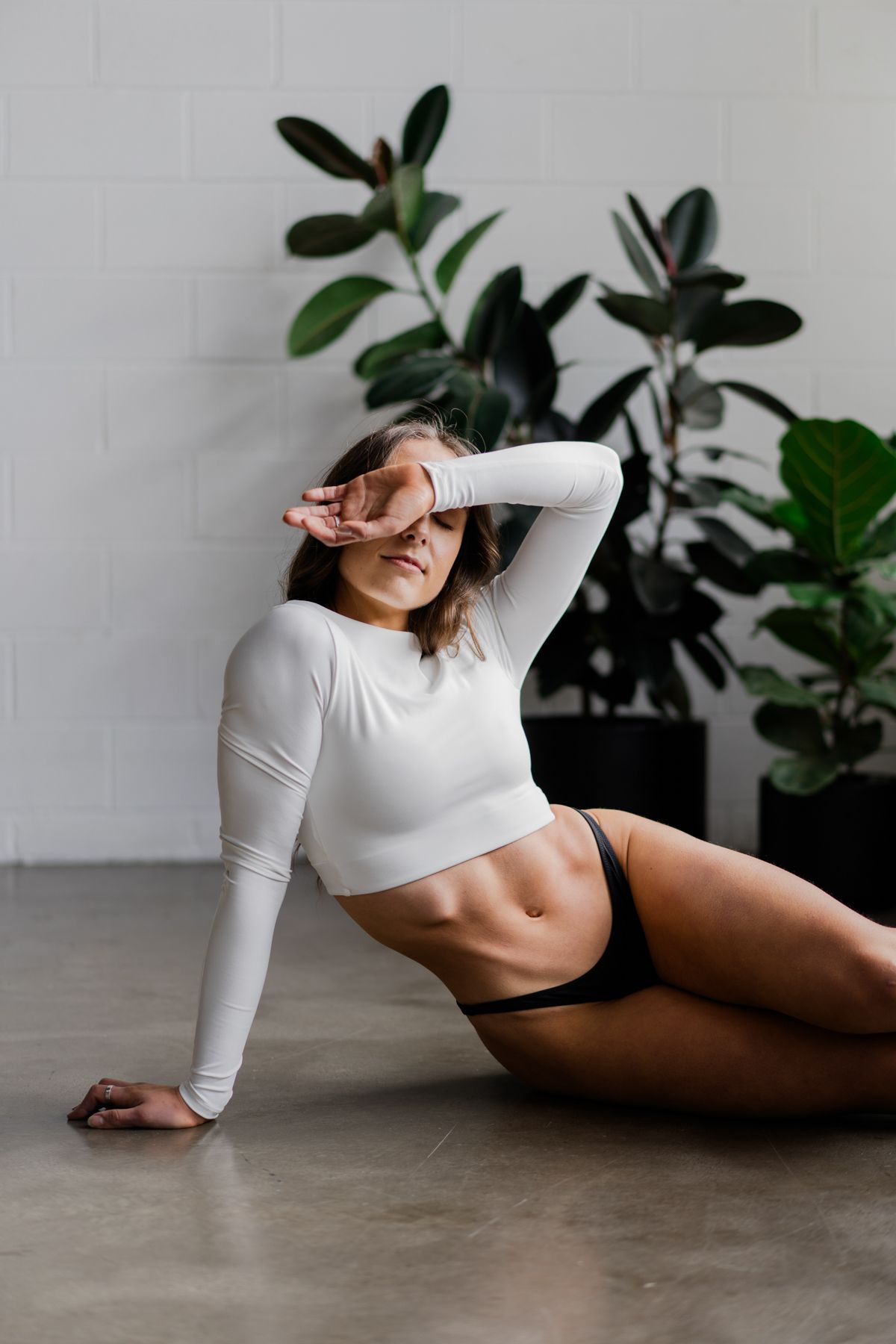 Fit woman in white crop top and black bikini bottoms sitting on the floor posing in front of green plants.