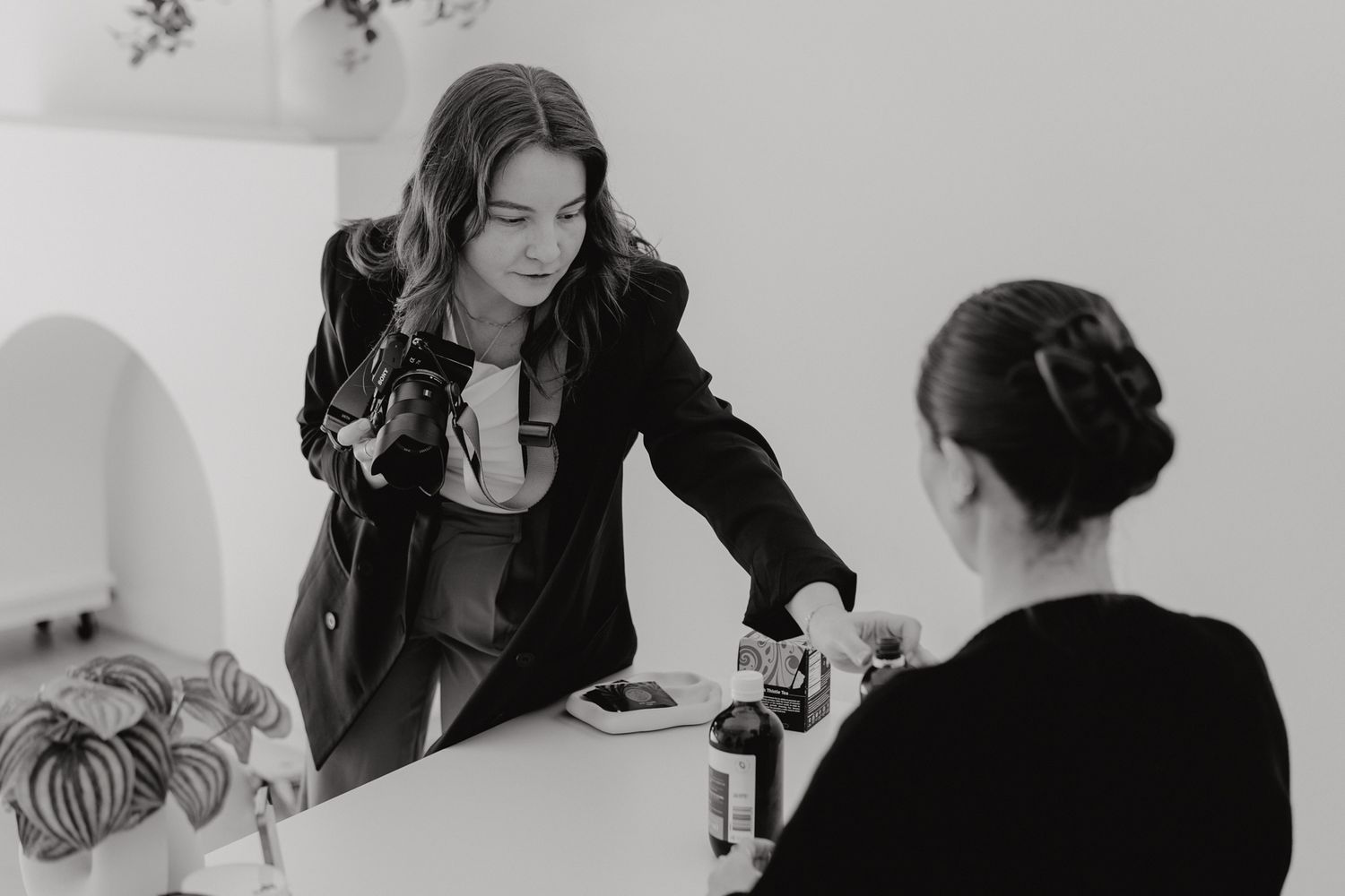 Female photographer assists client arranging products on white table in black and white studio photo.