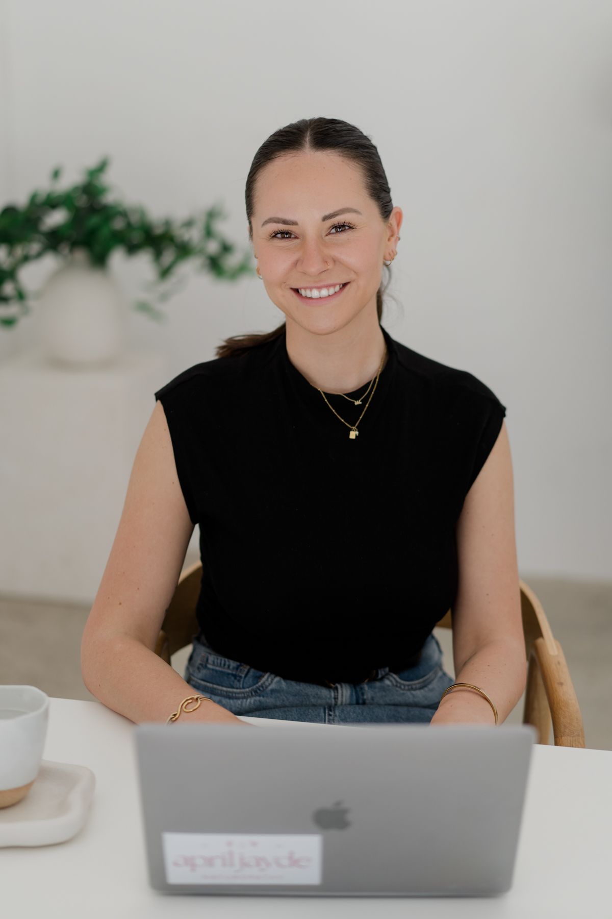 Smiling brunette woman in black top sitting at white desk with MacBook laptop.