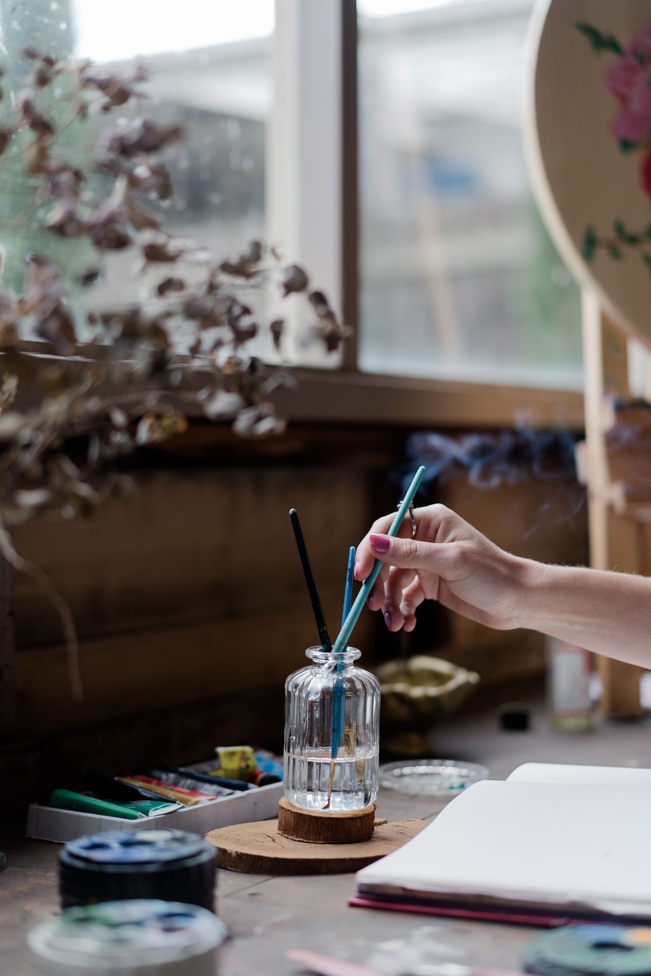 Close-up of artist hand selecting paintbrush from glass jar on a rustic wooden art studio desk.