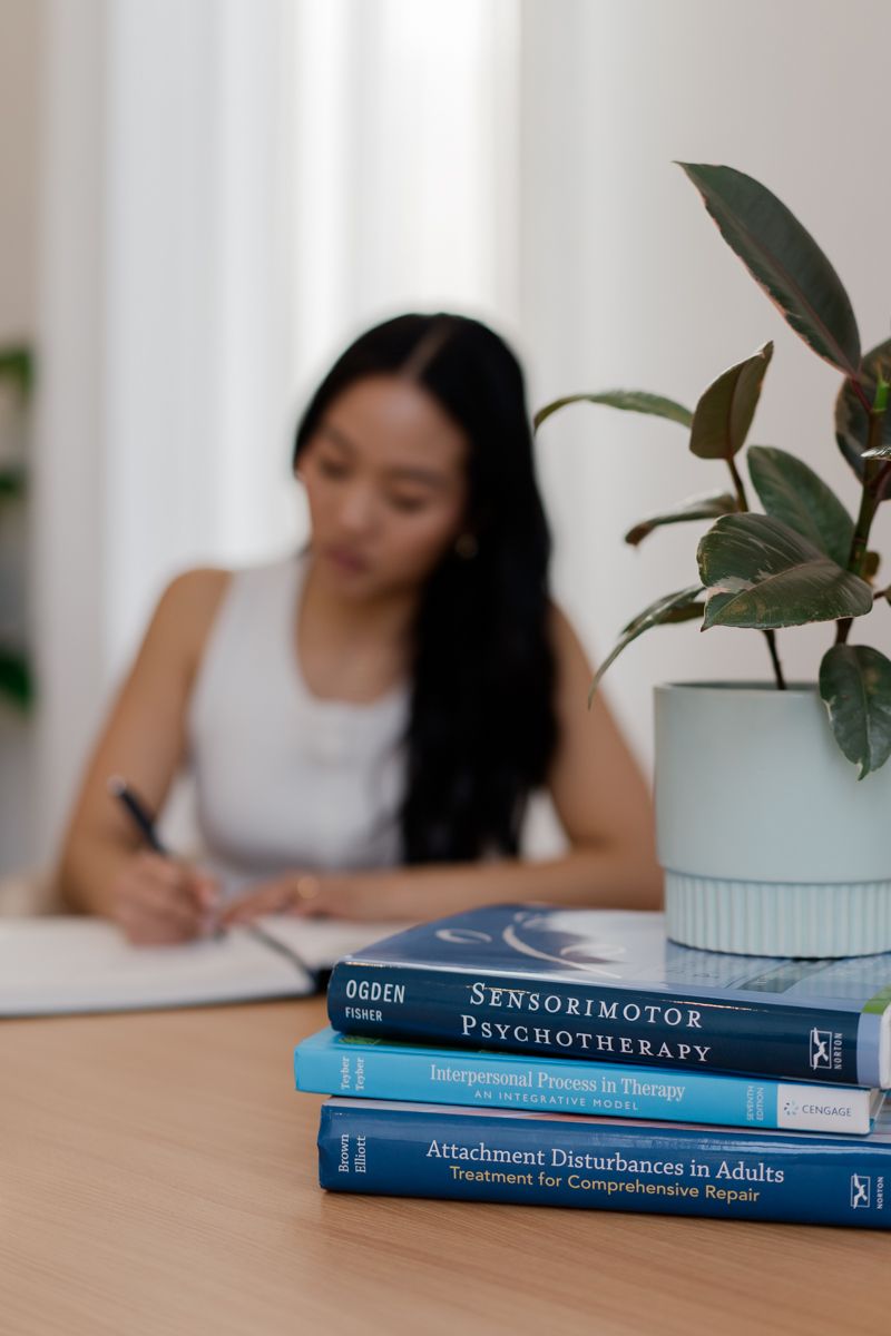 Psychology books and green plant in foreground with Asian woman writing at desk in background.