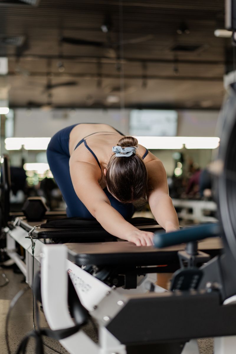 Woman in blue workout attire stretching forward on a Pilates reformer machine at a gym.