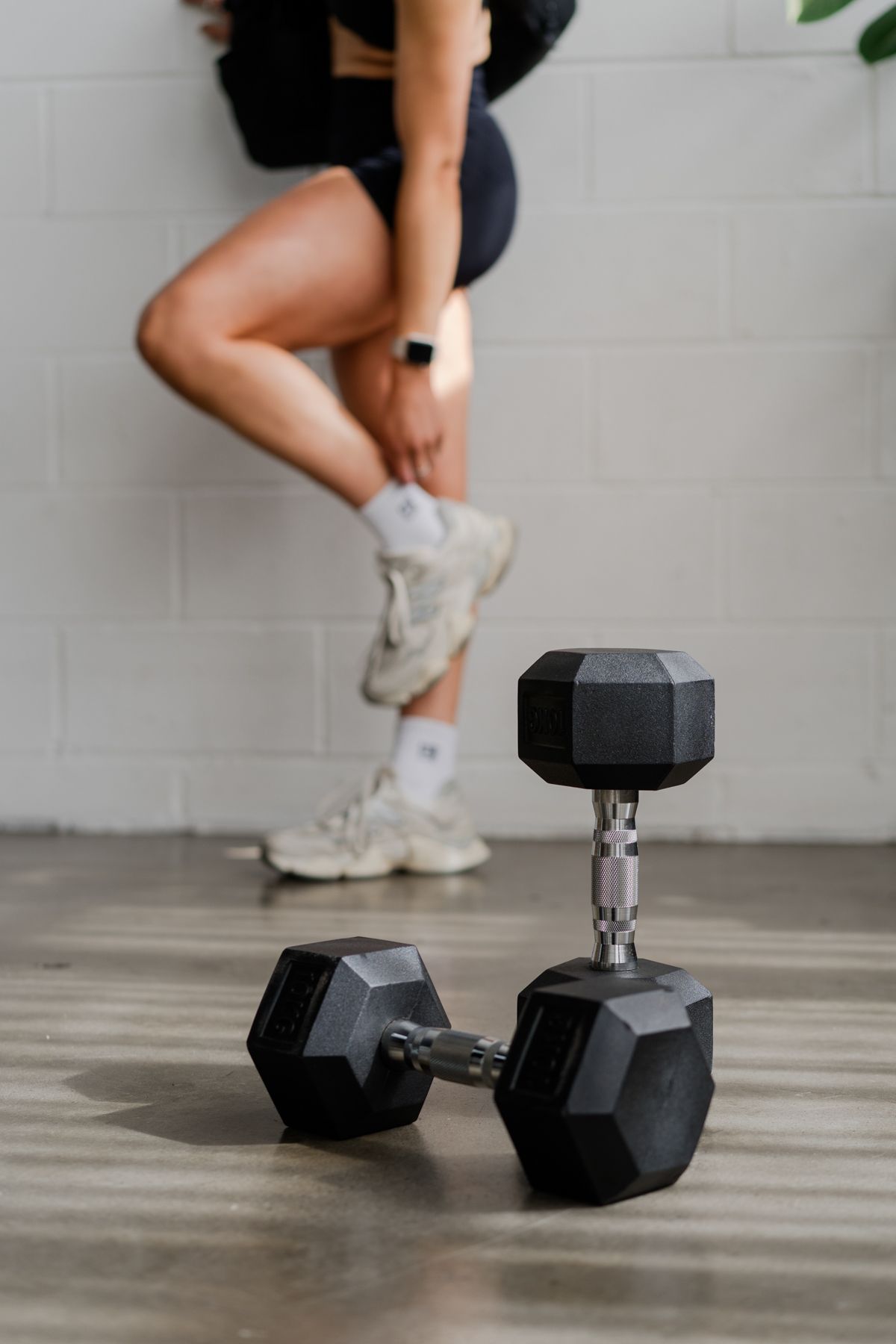 Woman in black workout clothes squatting near hex dumbbells on gym floor against white brick wall.