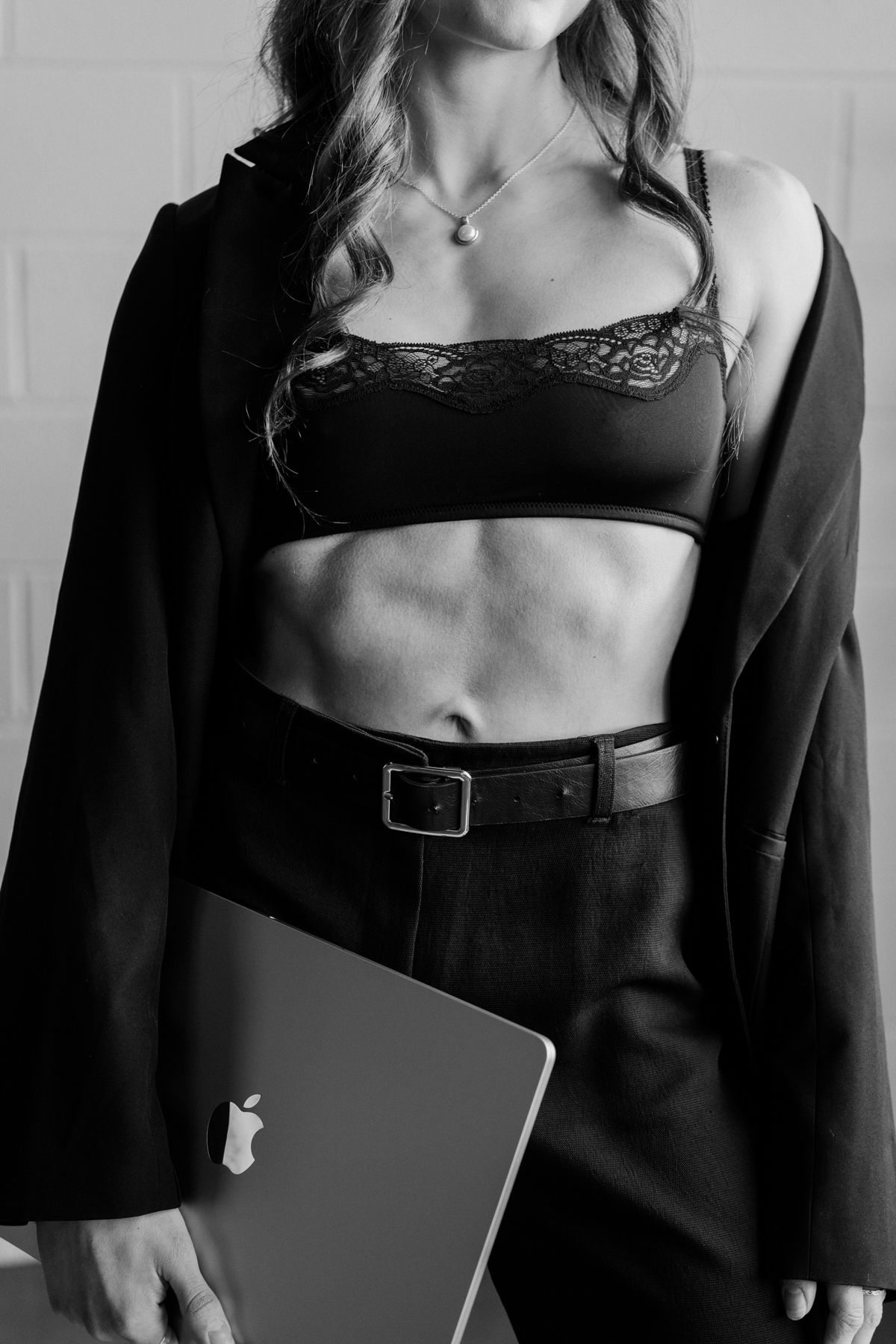 Black and white photo of woman in lacy bralette, blazer and trousers holding MacBook laptop.