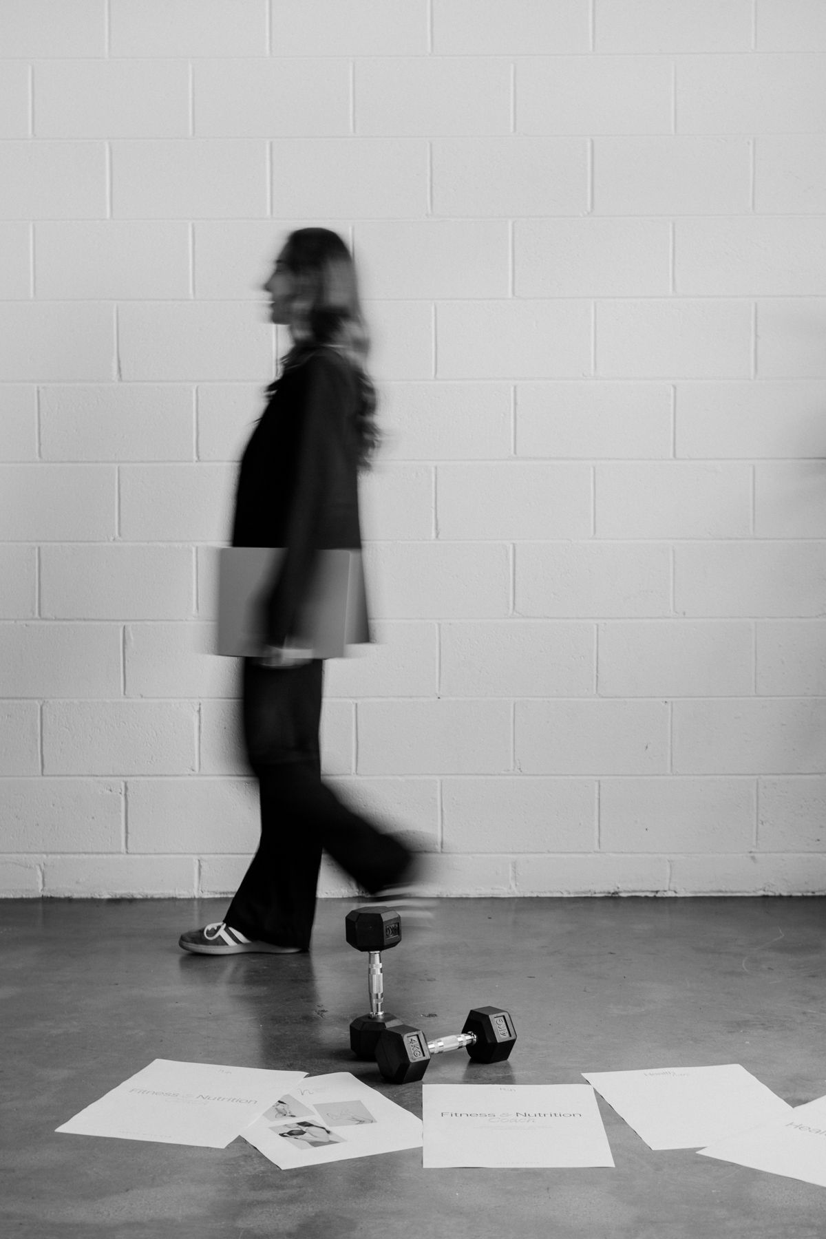 Black and white motion blur photo of woman walking past dumbbells and fitness coaching documents on studio floor.