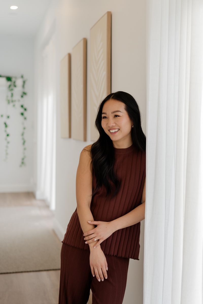 Smiling Asian woman in burgundy pleated outfit leans against white wall in bright hallway.