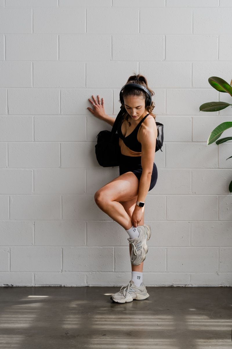 Fit woman in black athletic wear leans against white brick wall adjusting sneaker with headphones on.